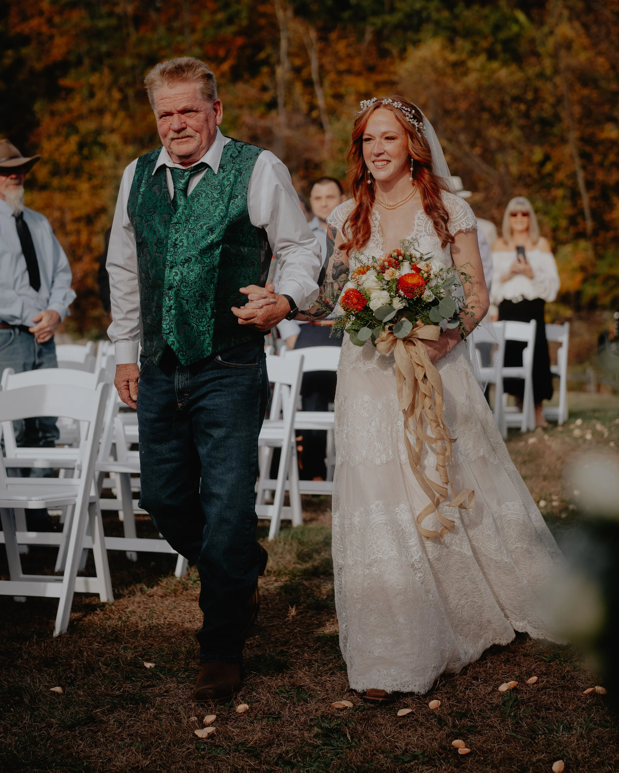 A bride walking down the aisle with her father at an outdoor wedding ceremony during fall, holding a bouquet of flowers.