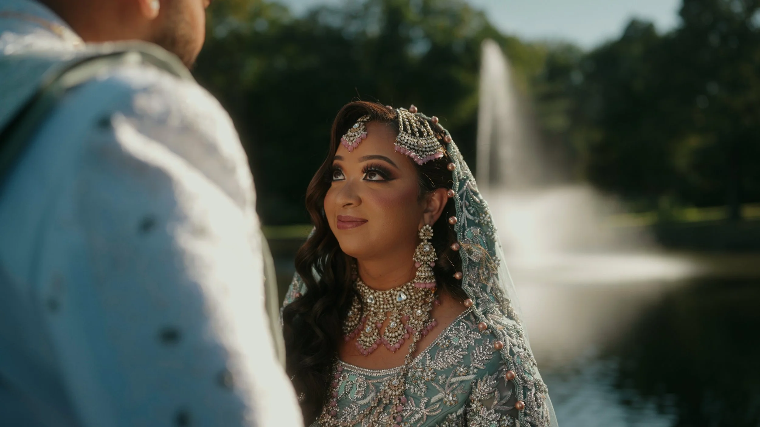 A woman dressed in traditional wedding attire, adorned with intricate jewelry, looks up at a man in a white embroidered outfit. They are outdoors near a fountain with water and trees in the background.