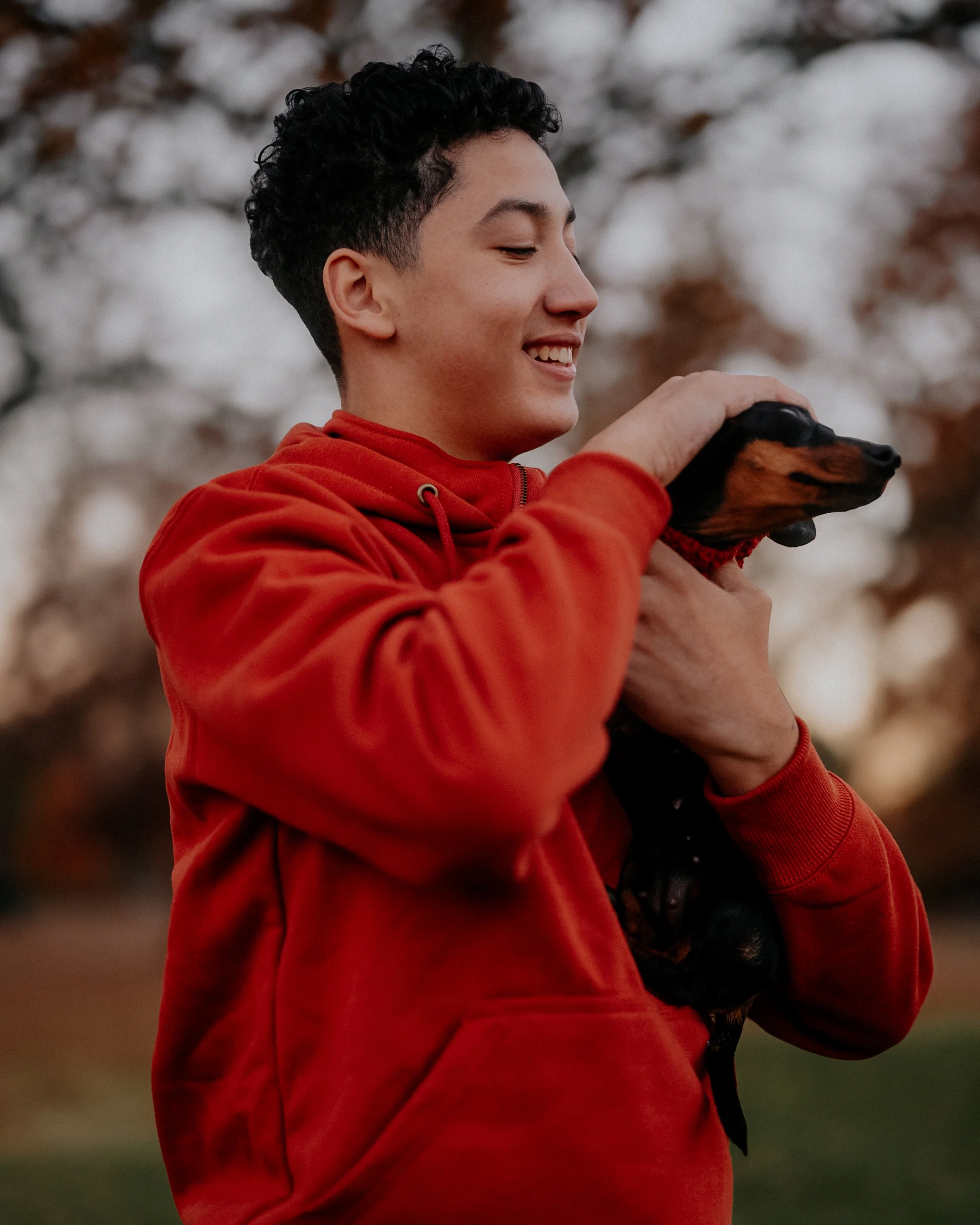 A young man with curly dark hair wearing a red hoodie, smiling, holding a small black and tan dog outdoors during autumn.