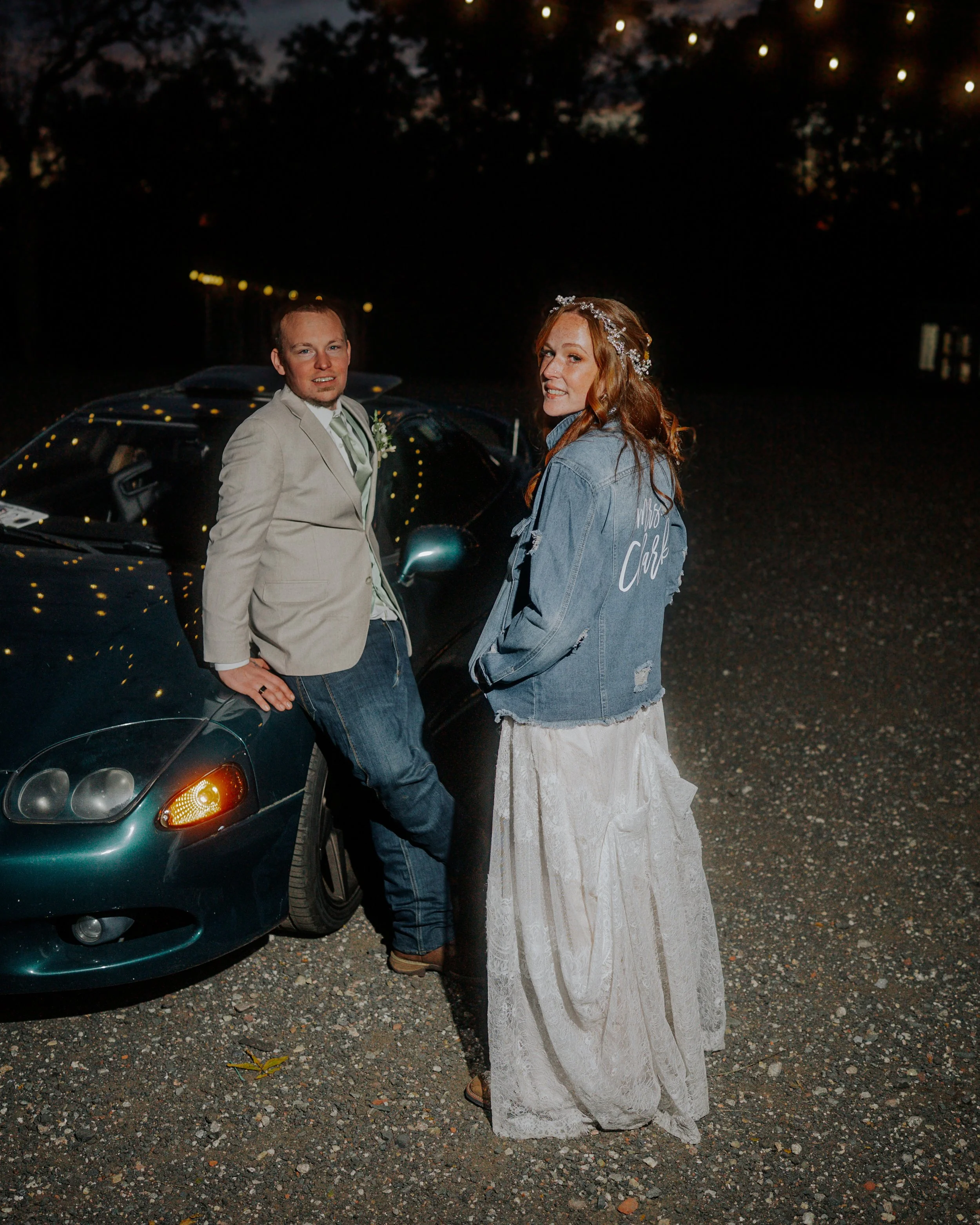 A man and a woman standing beside a black car outside at night, with trees in the background. The man is leaning on the car, dressed in a beige blazer and jeans, while the woman wears a long lace dress and a denim jacket with 'Mrs. Clark' written on 