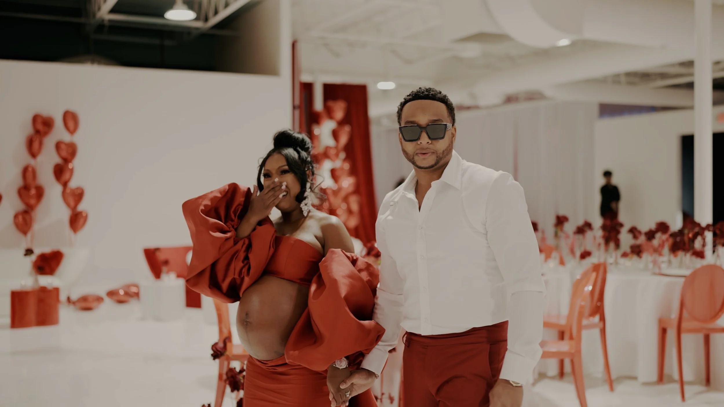 A pregnant woman in a red dress and large puffy sleeves, laughing and covering her mouth, is holding hands with a man wearing a white shirt, red pants, and sunglasses, in a decorated event space with heart-shaped balloons and red roses.