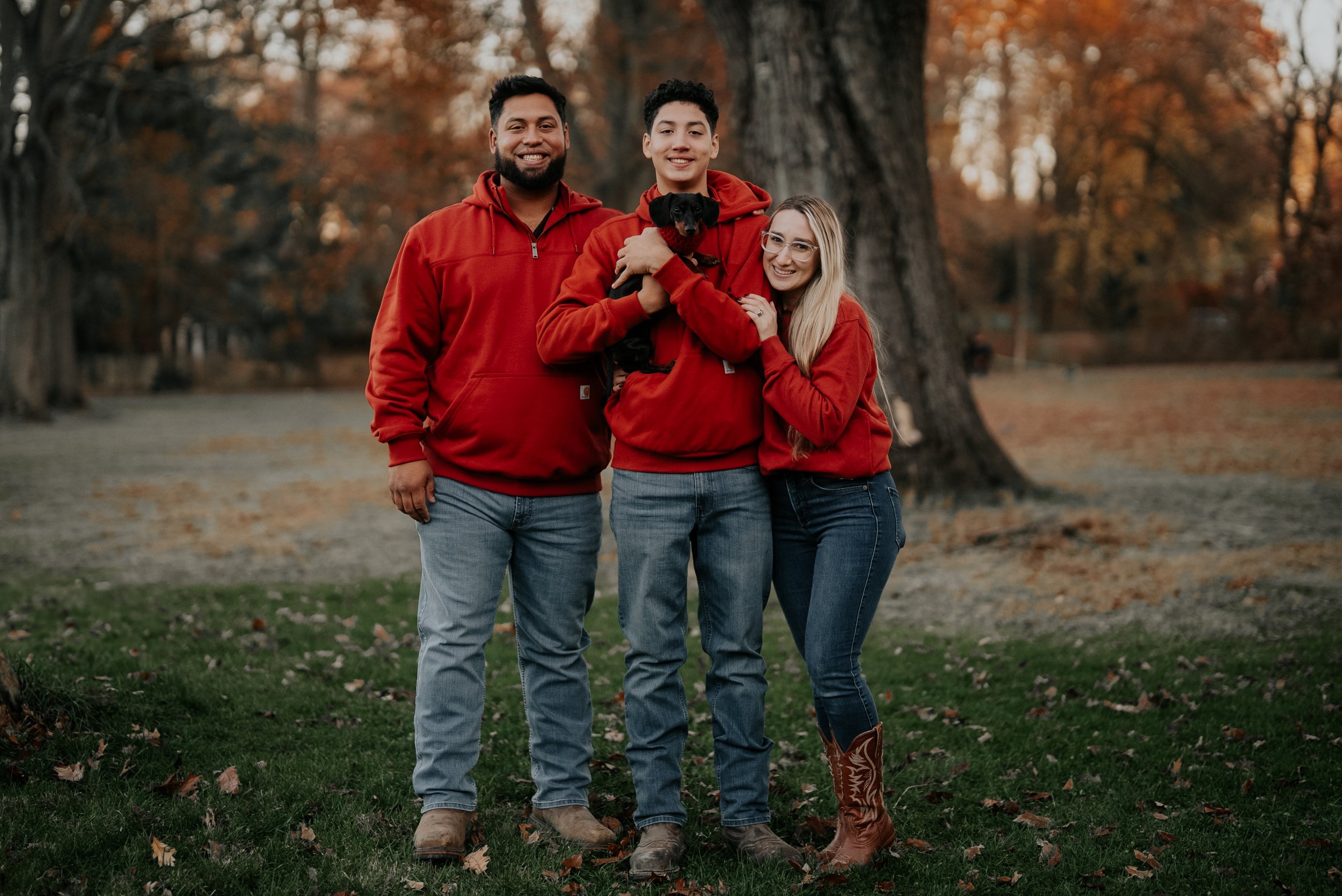 Three people in red hoodies standing together outdoors in a park during fall, with autumn leaves on the ground and trees in the background. One person is holding a small black dog.
