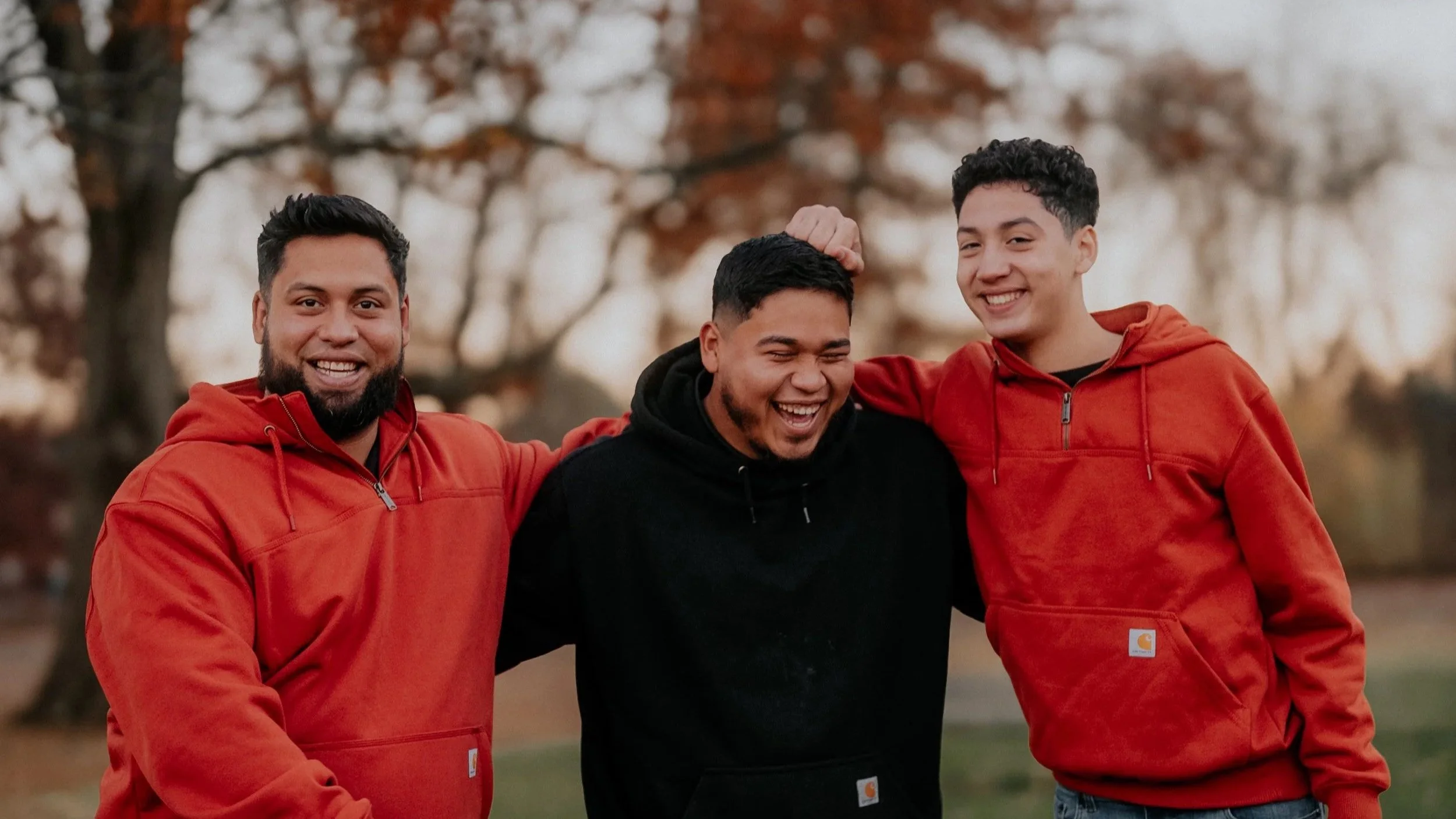 Three young men standing outdoors, smiling and laughing, with two of them wearing red hoodies and the middle wearing a black hoodie, with autumn trees in the background.
