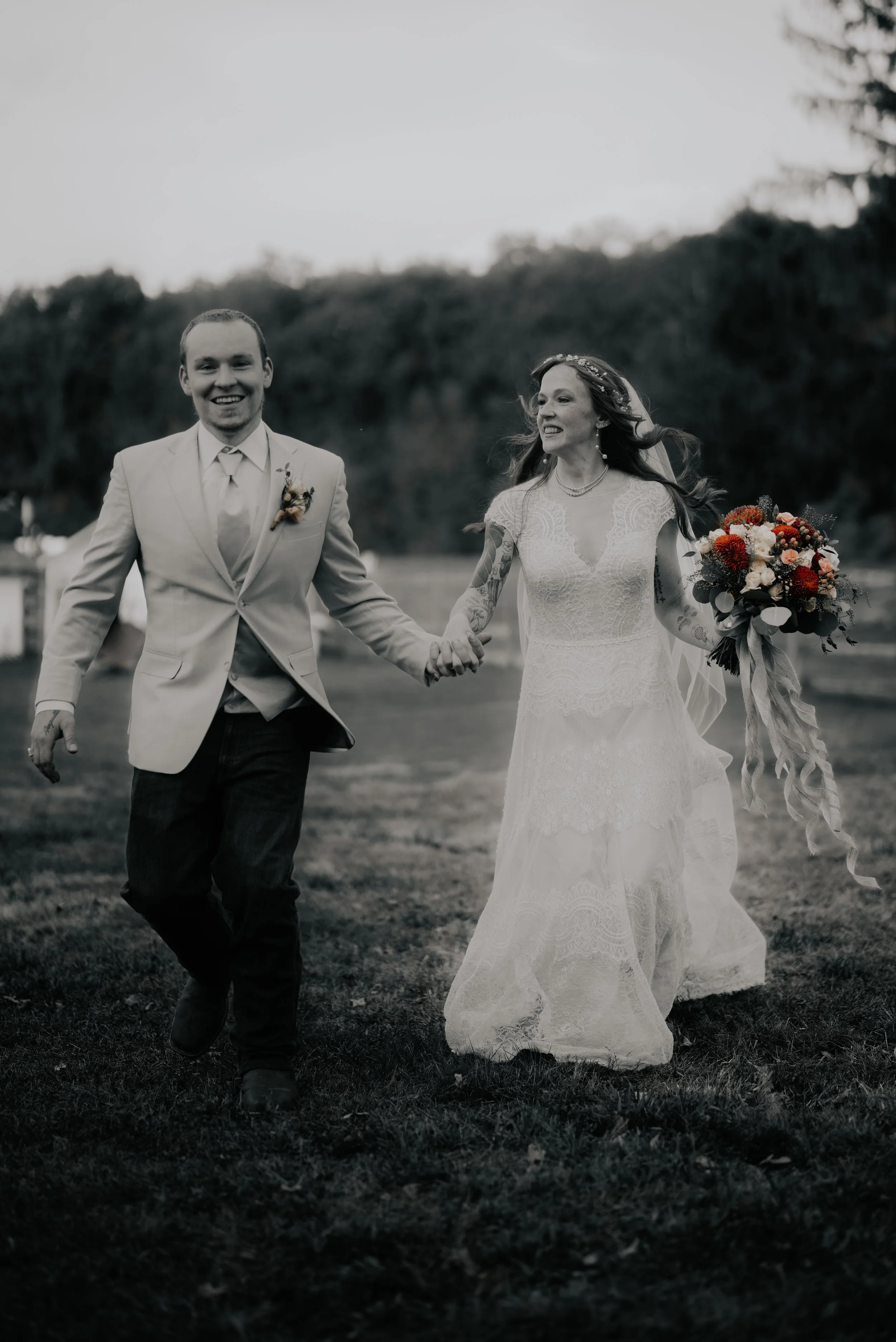 A black and white photo of a smiling couple holding hands, walking across a grassy field, with the bride holding a colorful bouquet of flowers.