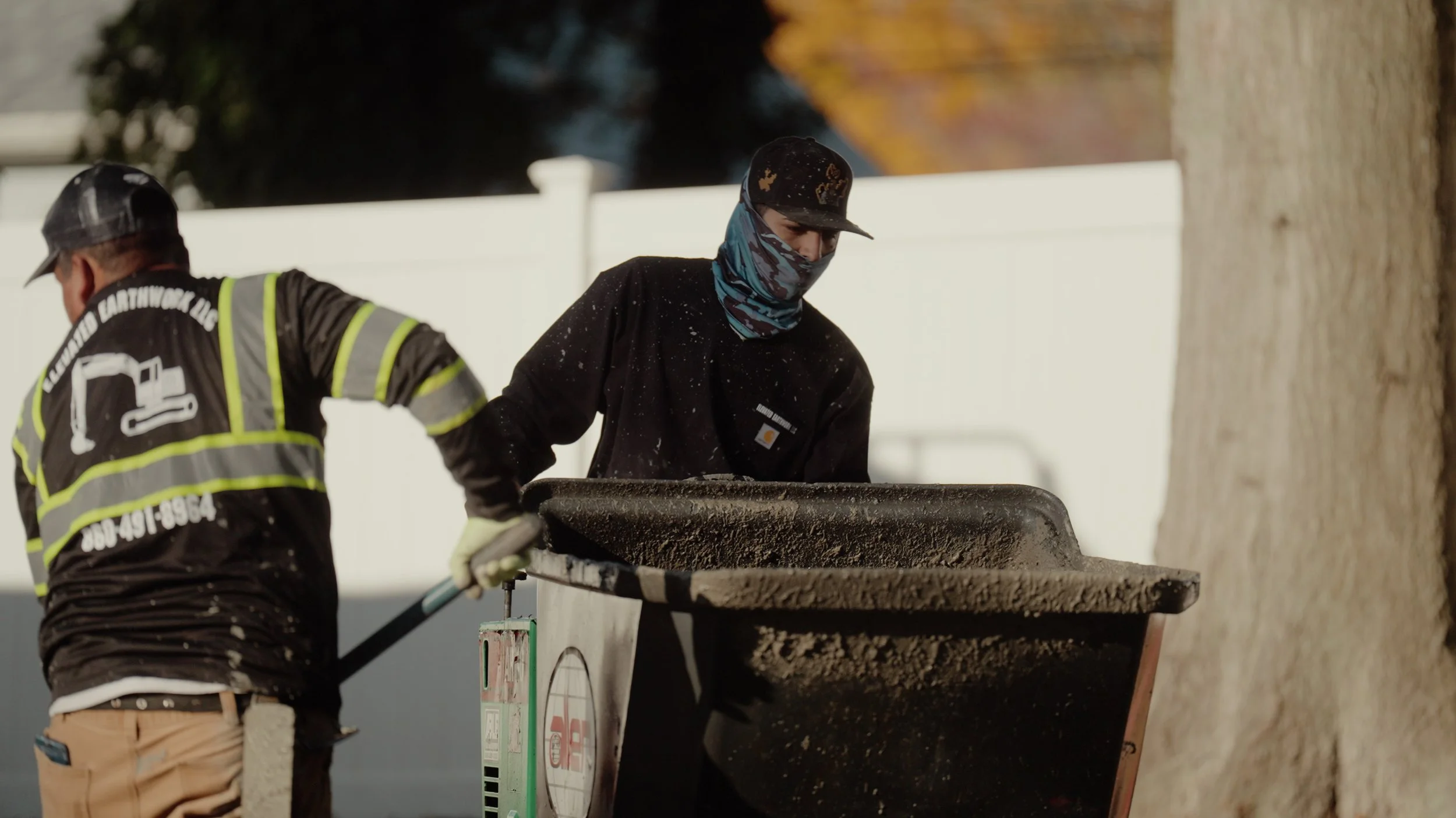 Two workers, one in a black jacket and a mask, shoveling dirt into a wheelbarrow with a tree in the background.