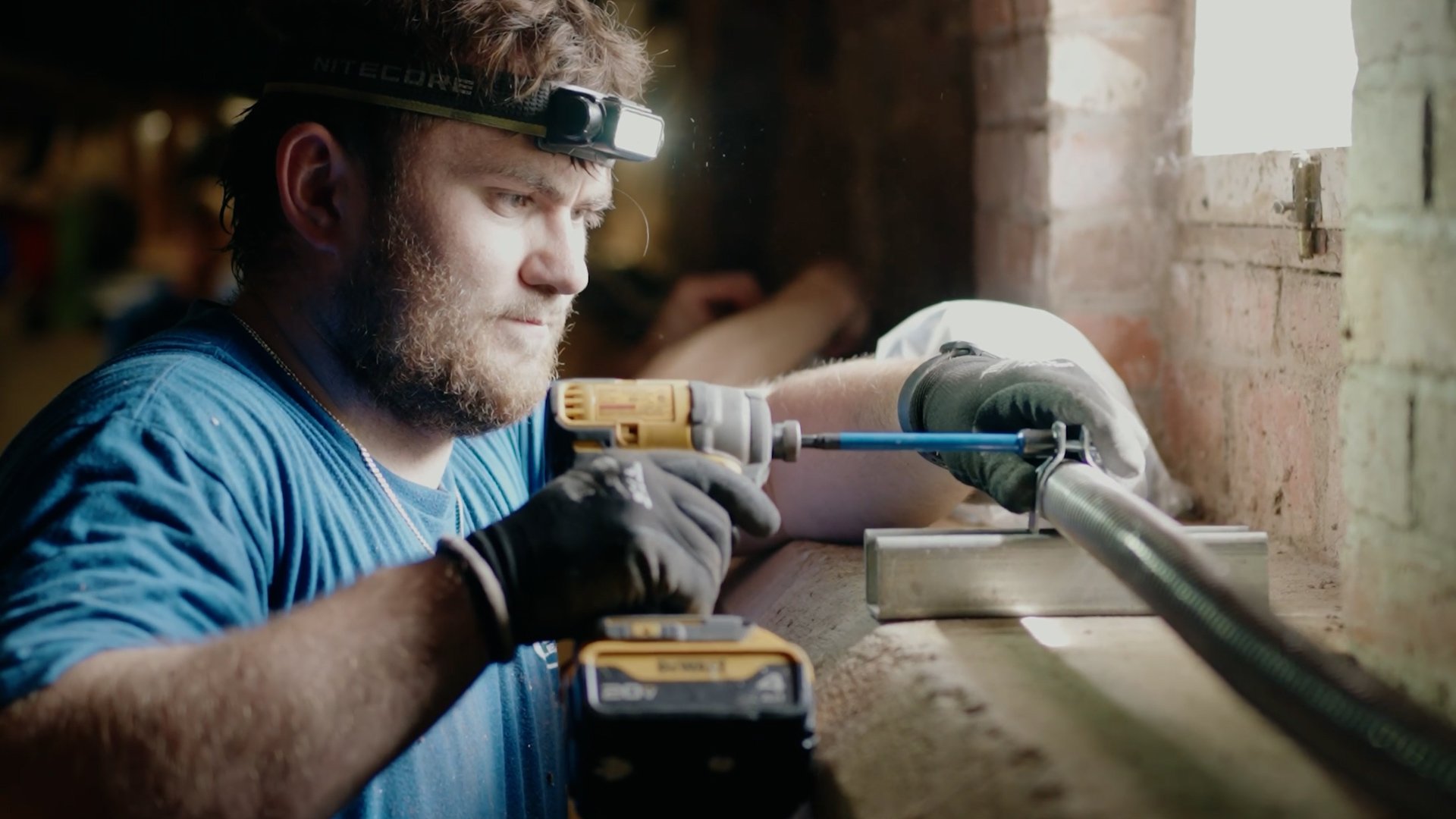 A man with a headlamp, beard, and blue shirt uses a cordless drill to work on a metal pipe in a workshop with brick walls and natural light coming through a window.