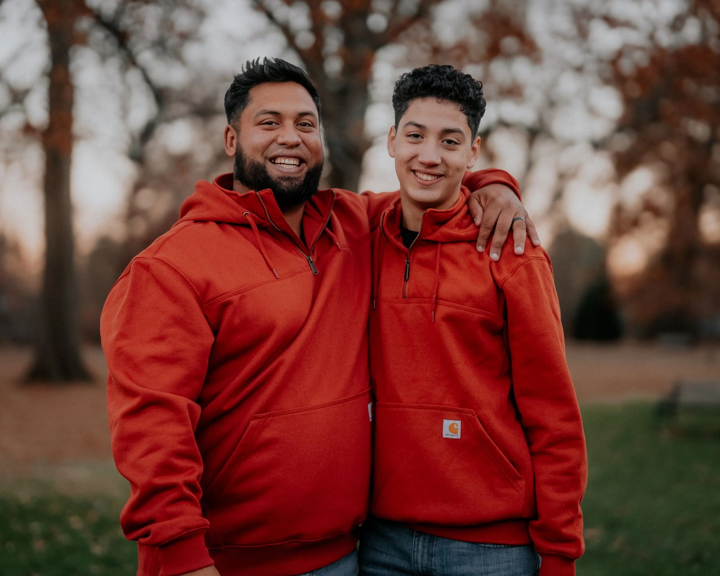 Two men smiling, standing outdoors in a park with autumn trees in the background, wearing matching red hoodies, with the younger man's arm around the older man's shoulder.