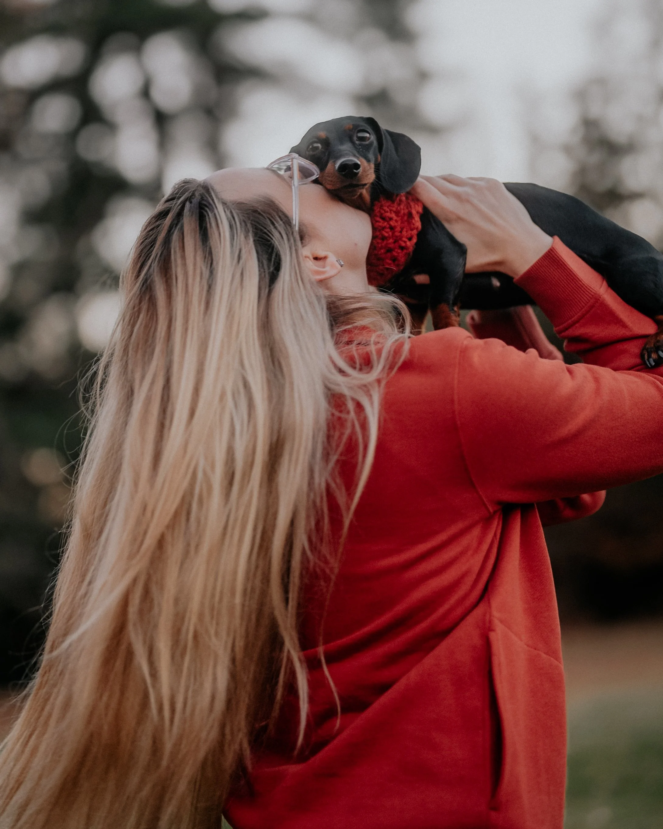 A woman with long blonde hair and glasses holding a small black and tan Dachshund dog with a red scarf outdoors.