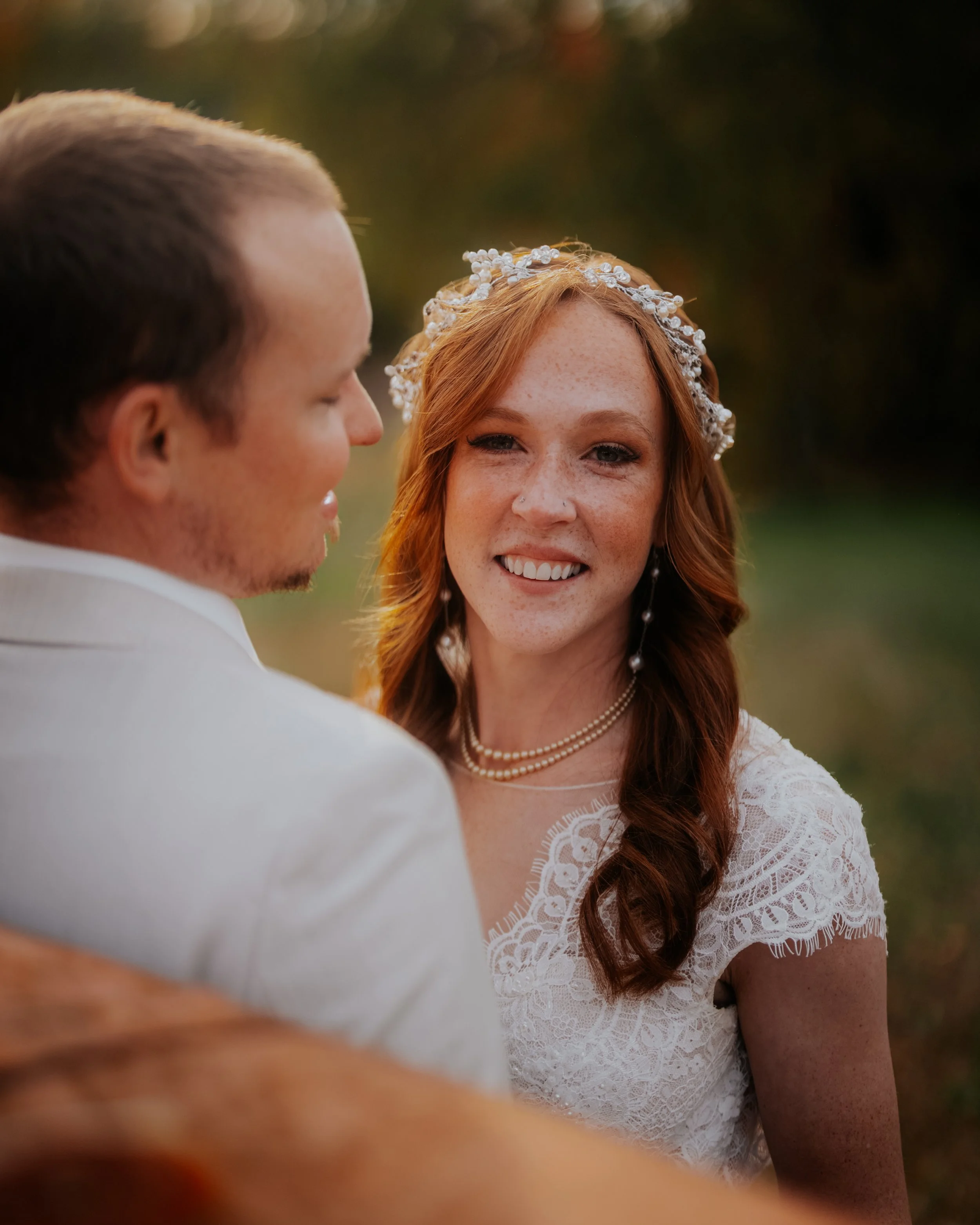 A woman with red hair, freckles, wearing a white lace dress, pearl necklace, and floral headband, smiling at a man with short brown hair, in a white suit, outdoors at sunset.