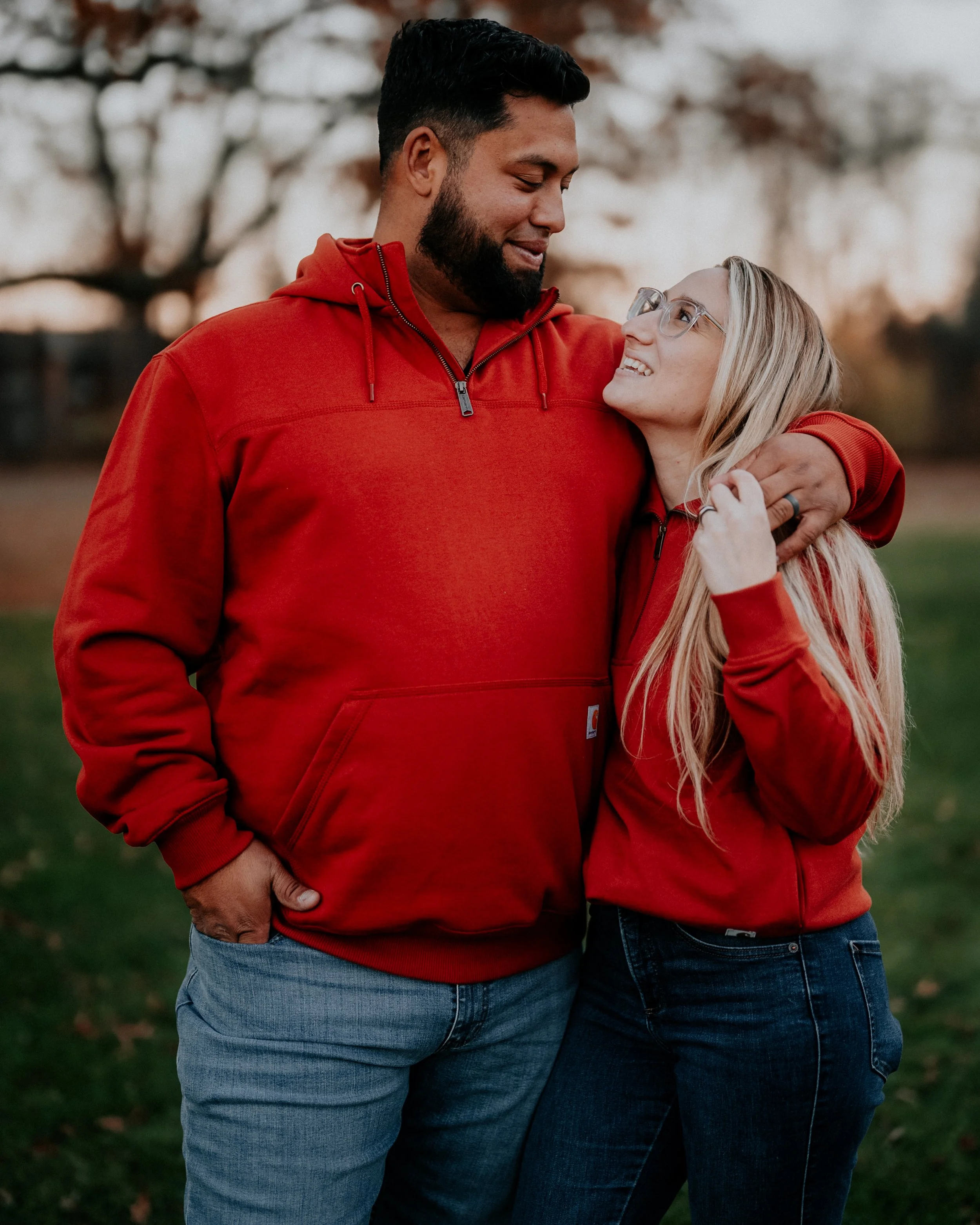 A man and woman stand close together outdoors during sunset, smiling and looking at each other. They are wearing red hoodies, with the man having his arm around the woman, and the woman looking up at the man.
