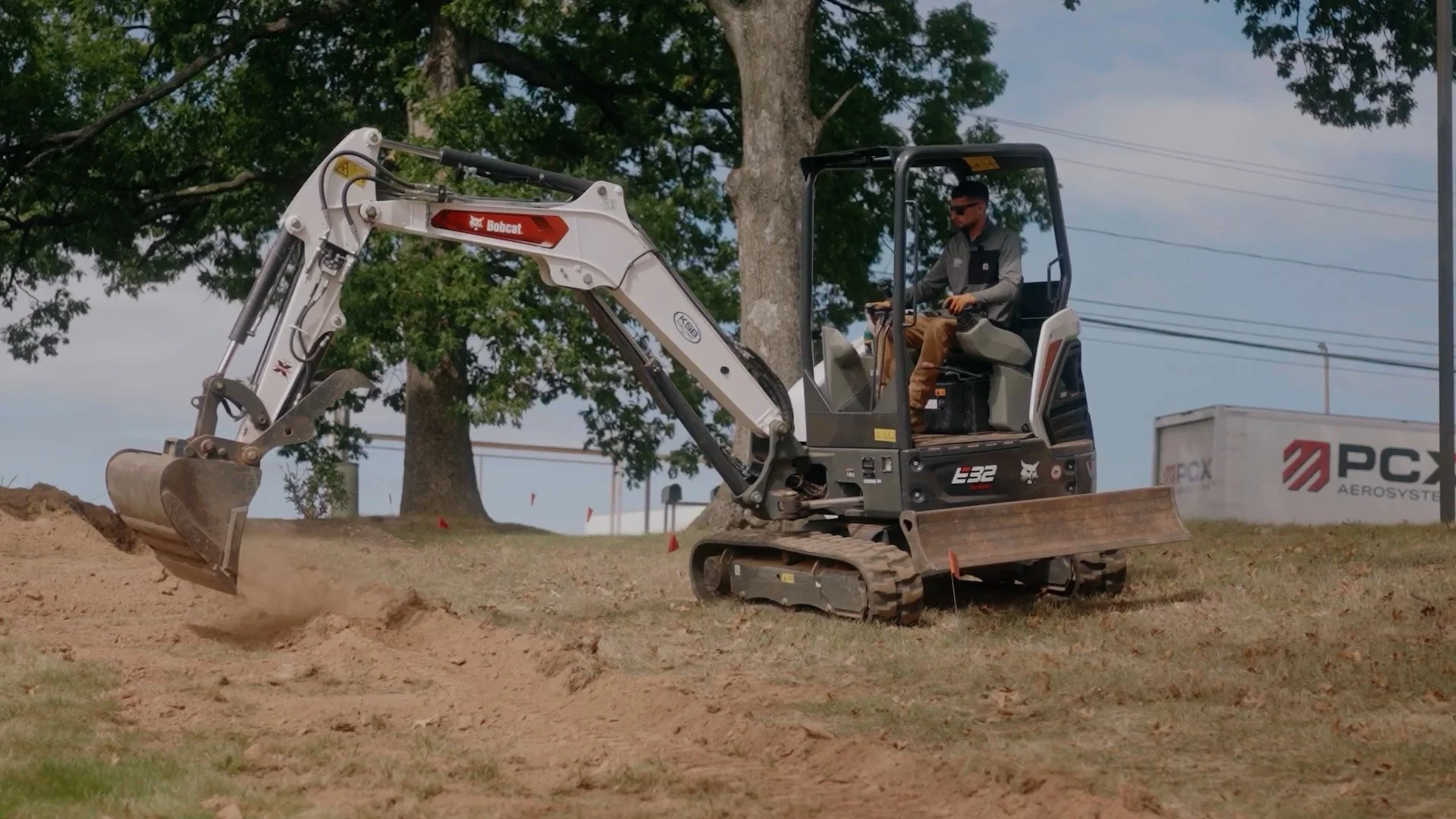 A person operating a Bobcat mini excavator on a patch of dirt with a tree, power lines, and a PCS Aerosystems sign in the background.