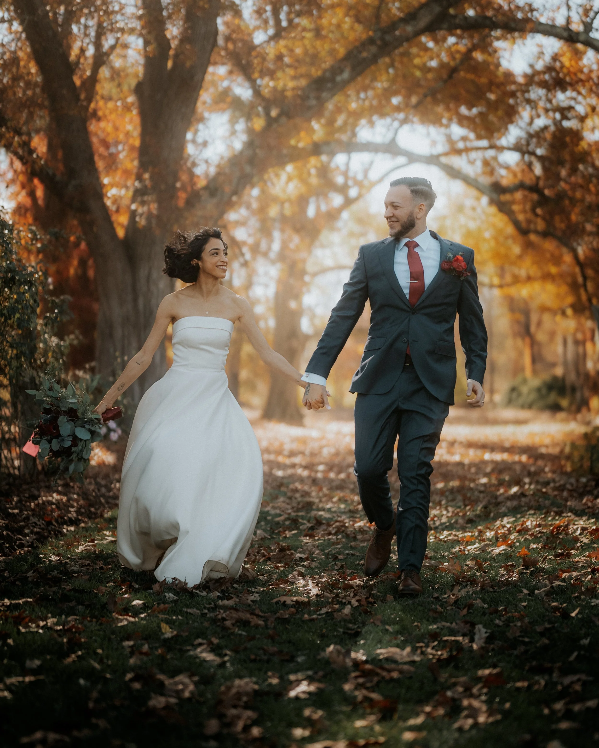 A bride and groom walking hand in hand through a fall forest, smiling at each other.