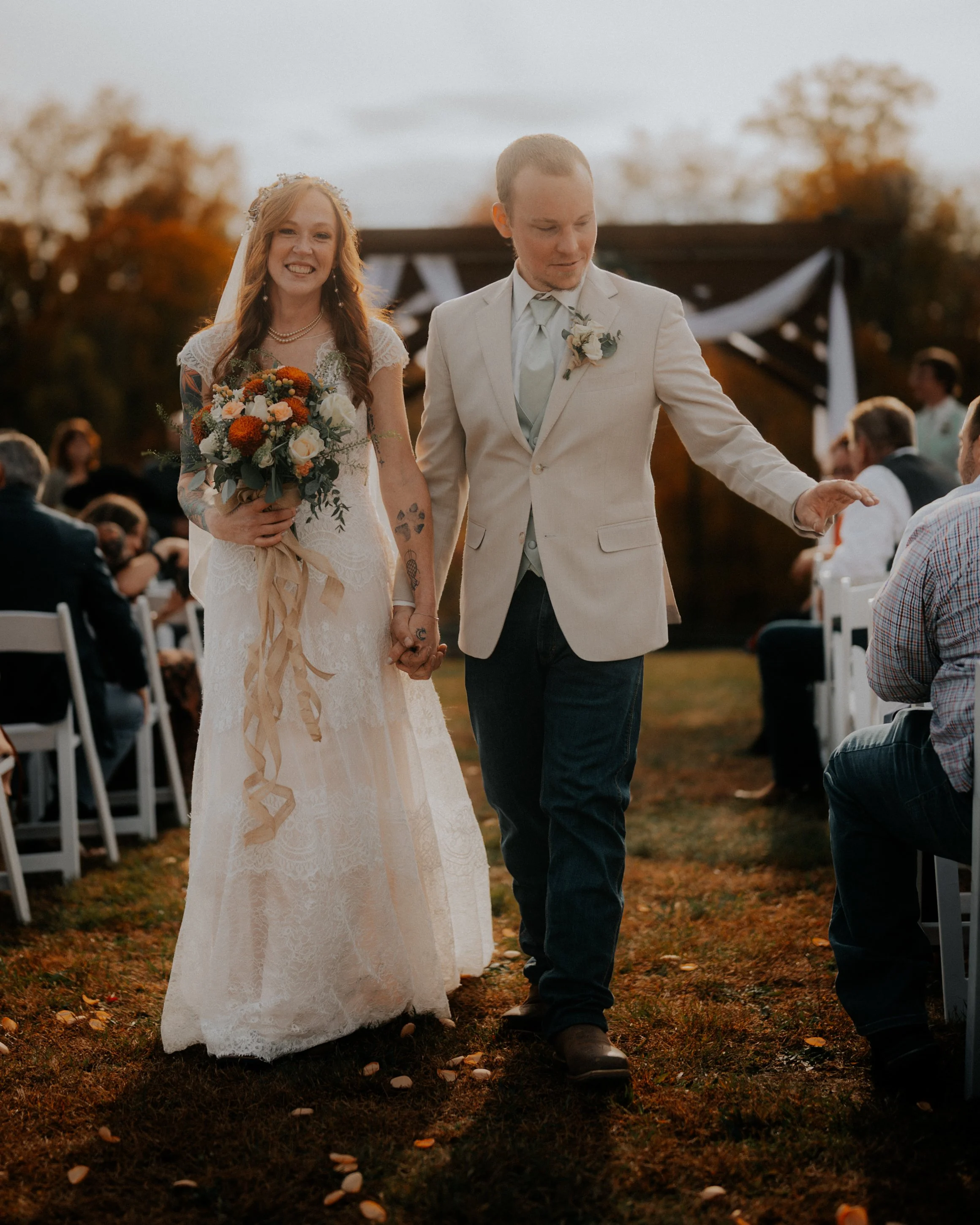 A newlywed couple walking hand-in-hand outdoors during their wedding ceremony, with guests seated on both sides, a rustic backdrop, and soft, warm lighting.