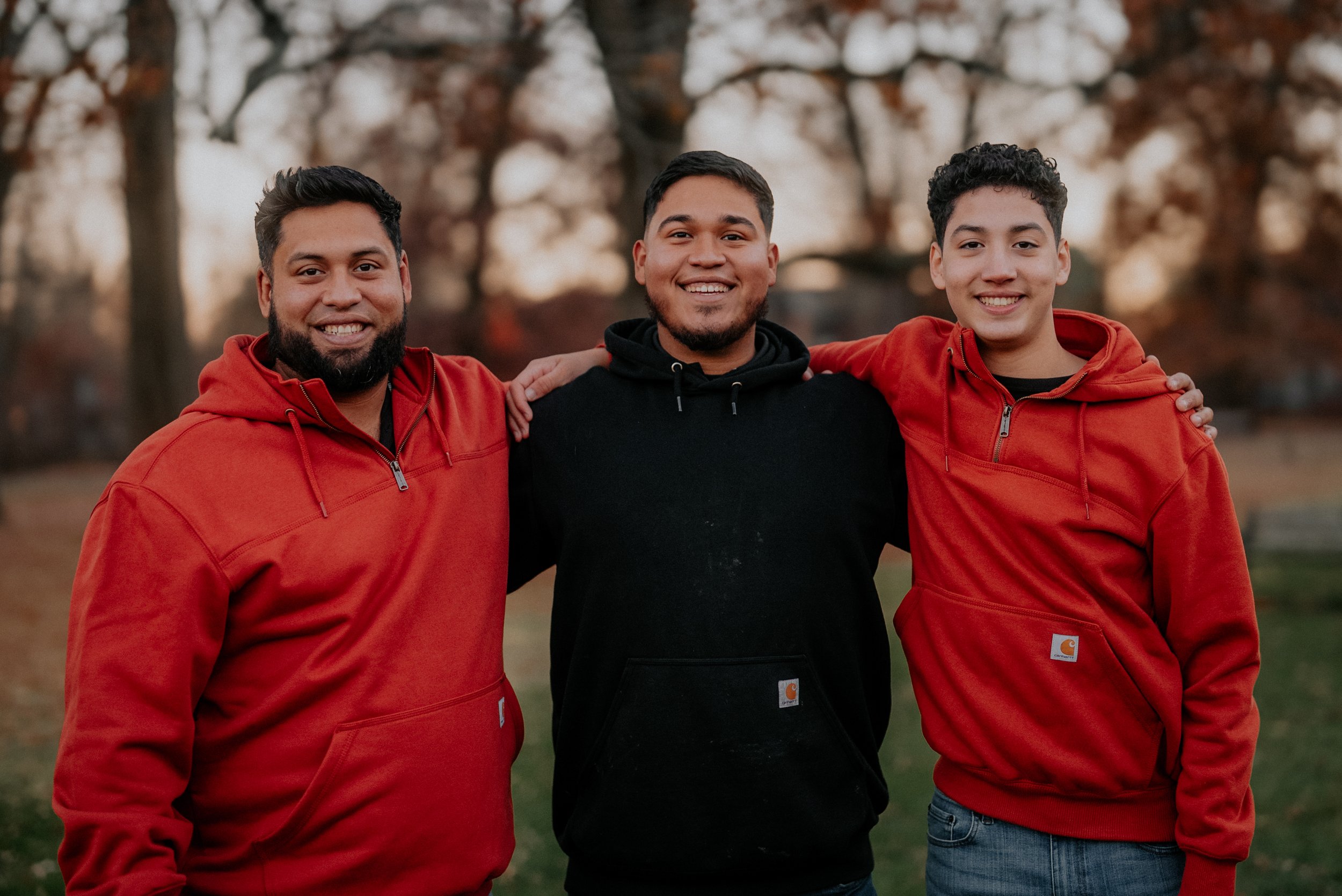 Three friends standing outdoors with their arms around each other's shoulders during autumn, smiling at the camera. Two are wearing red hoodies and the one in the middle is wearing a black hoodie.