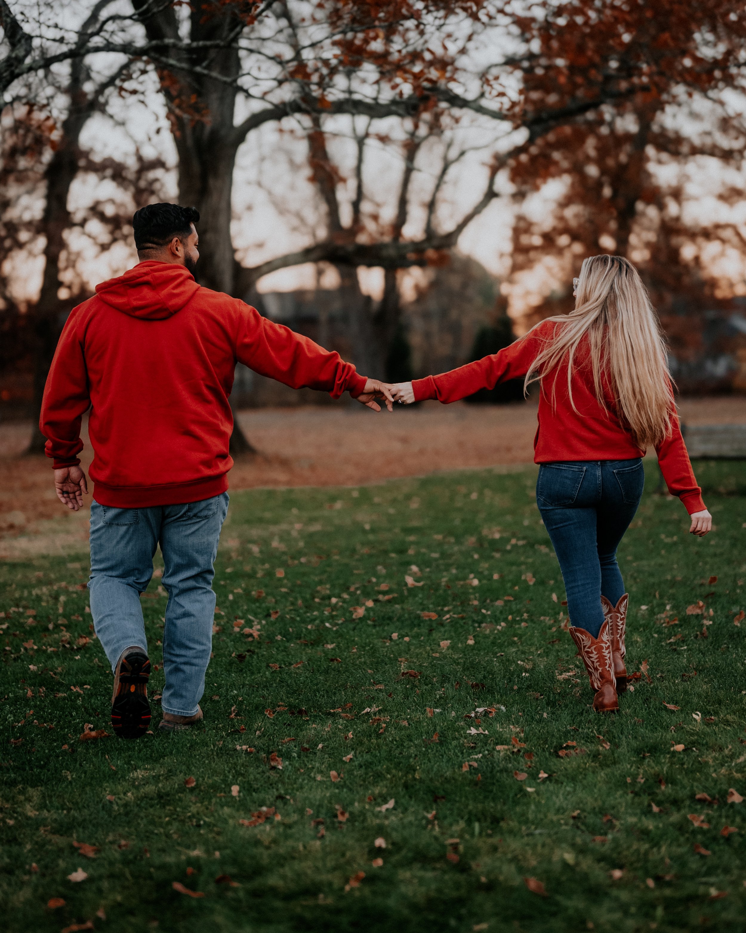 A couple walking hand in hand in a park during autumn, both wearing red hoodies, with fall trees in the background