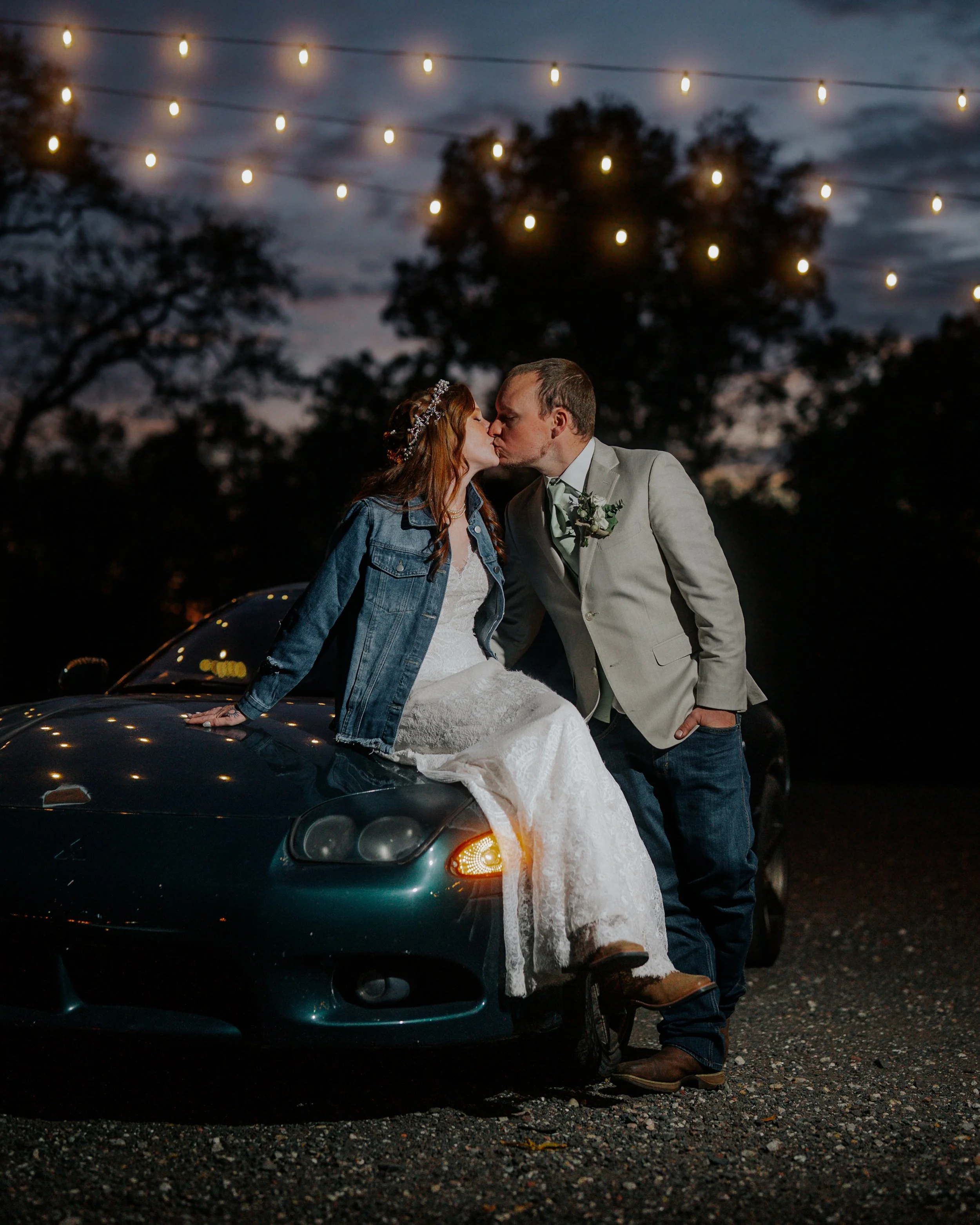 A couple in wedding attire sharing a kiss at night, with string lights overhead and trees in the background. One person is sitting on the hood of a dark-colored car, while the other is standing beside them.