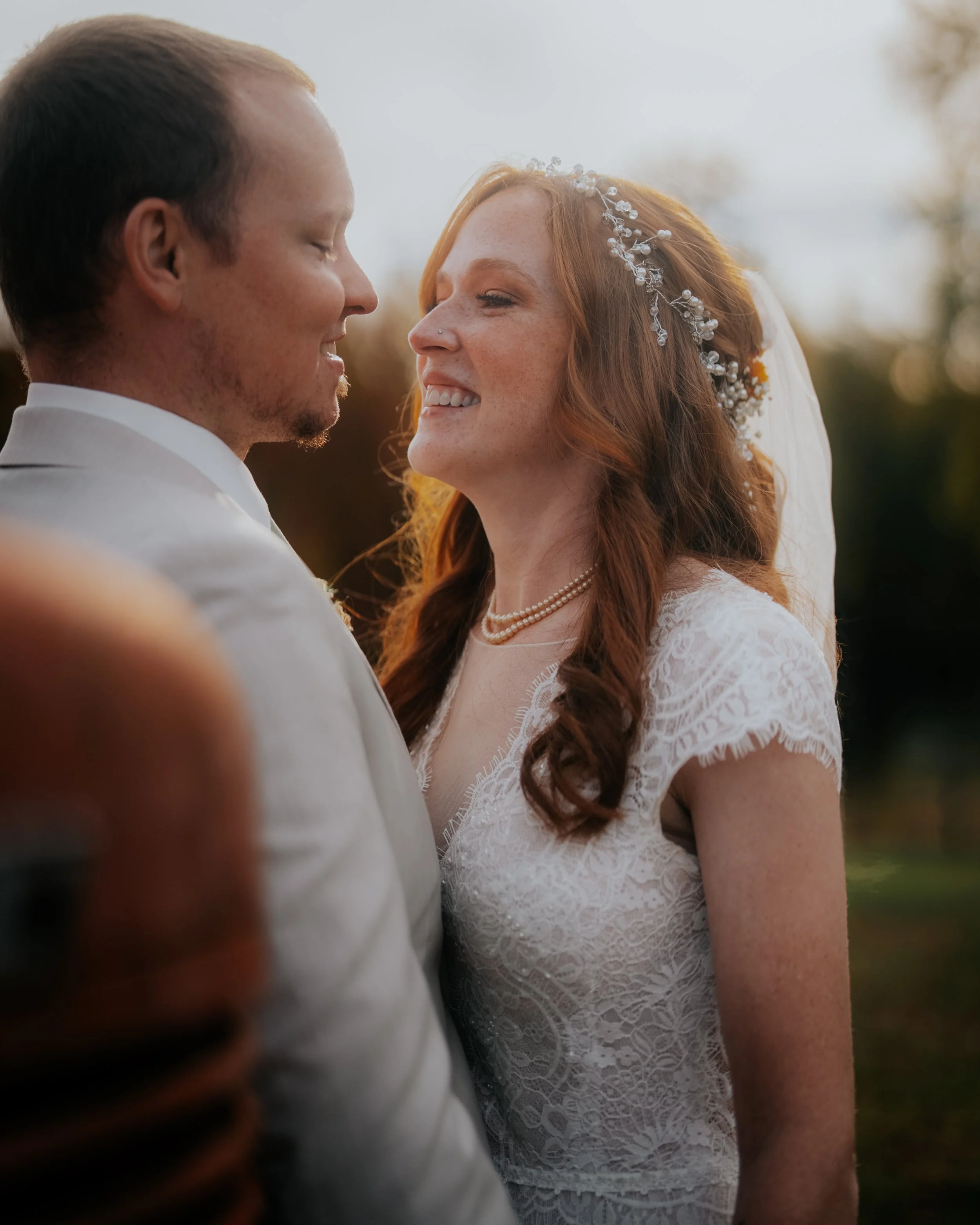A bride and groom standing close together outdoors at sunset, smiling and about to kiss, with a blurred natural background.