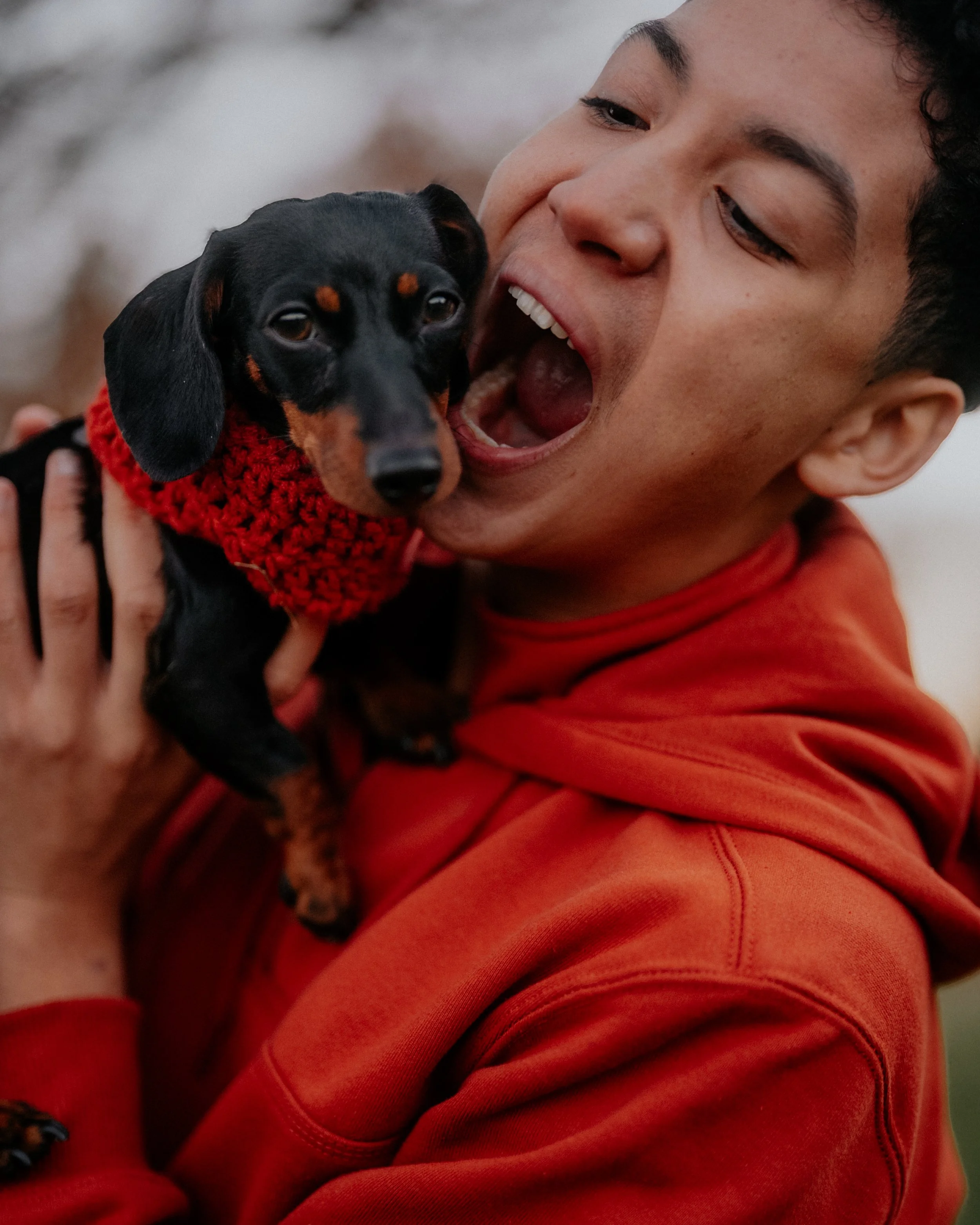 A young man with short dark curly hair smiling and holding a small black and tan Dachshund puppy wearing a red knit sweater close to his face outdoors.