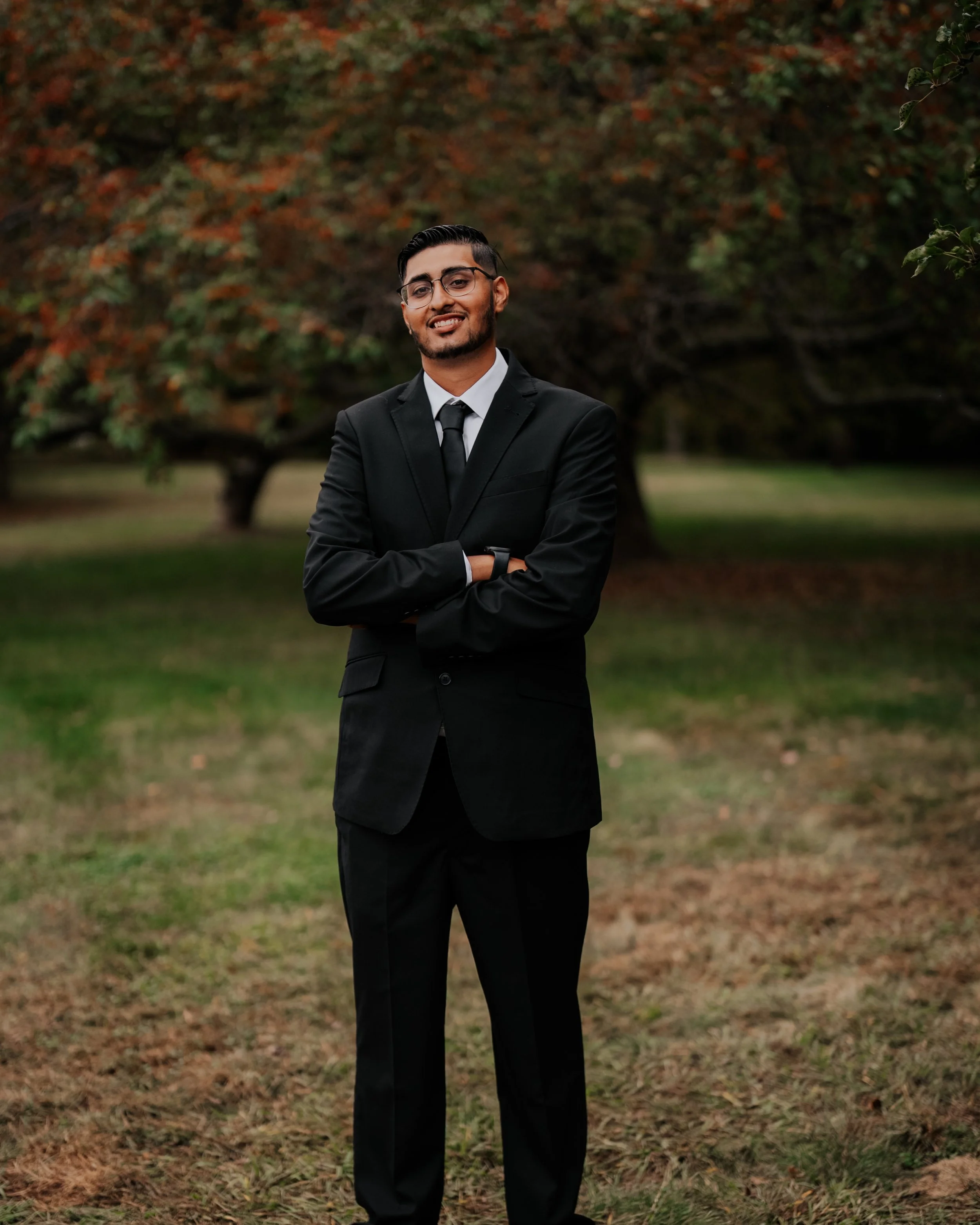 A young man in a black suit, white shirt, and black tie standing outdoors with arms crossed, in front of a backdrop of trees with autumn-colored leaves.