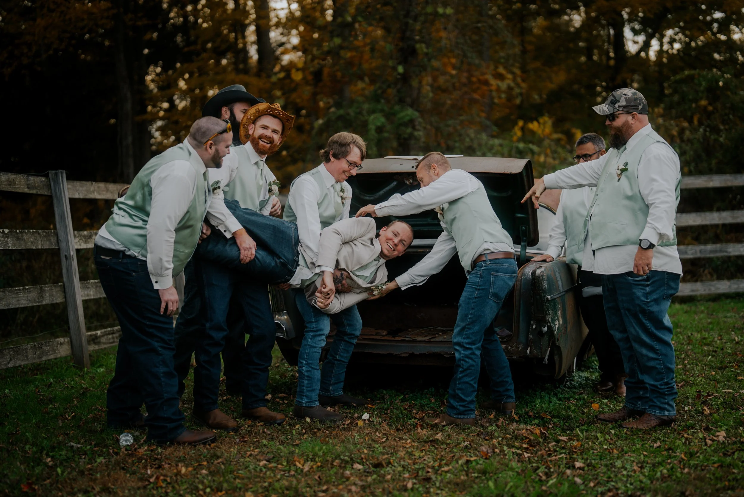 A group of men dressed in matching light-colored vests and shirts are gathered around an open trunk of a vintage car in an outdoor setting with fall foliage. They are all smiling and appear to be having fun, with some helping a man dressed in a light