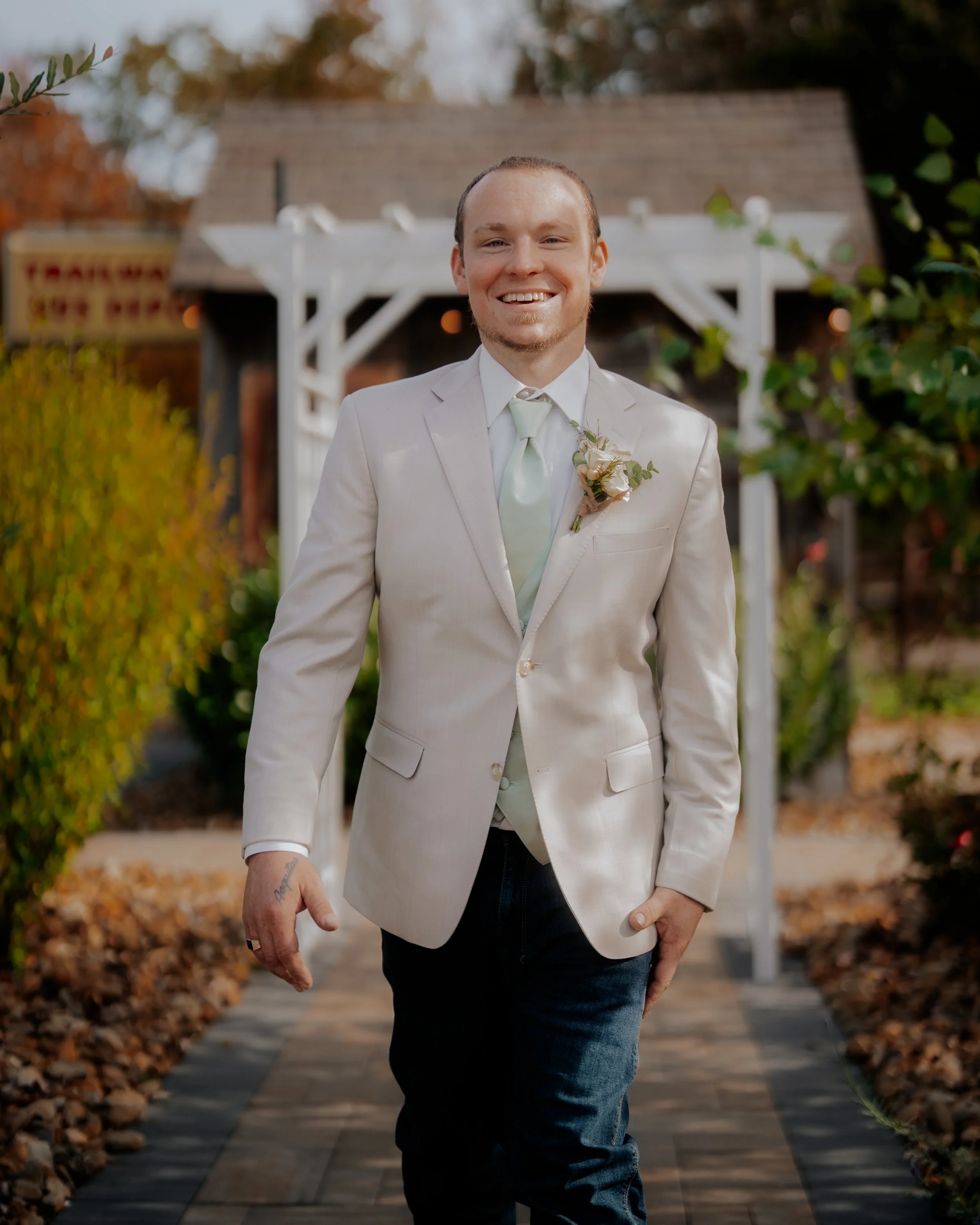 A smiling man in a light-colored suit walking outside on a pathway with autumn leaves, a white arch structure, and greenery in the background.
