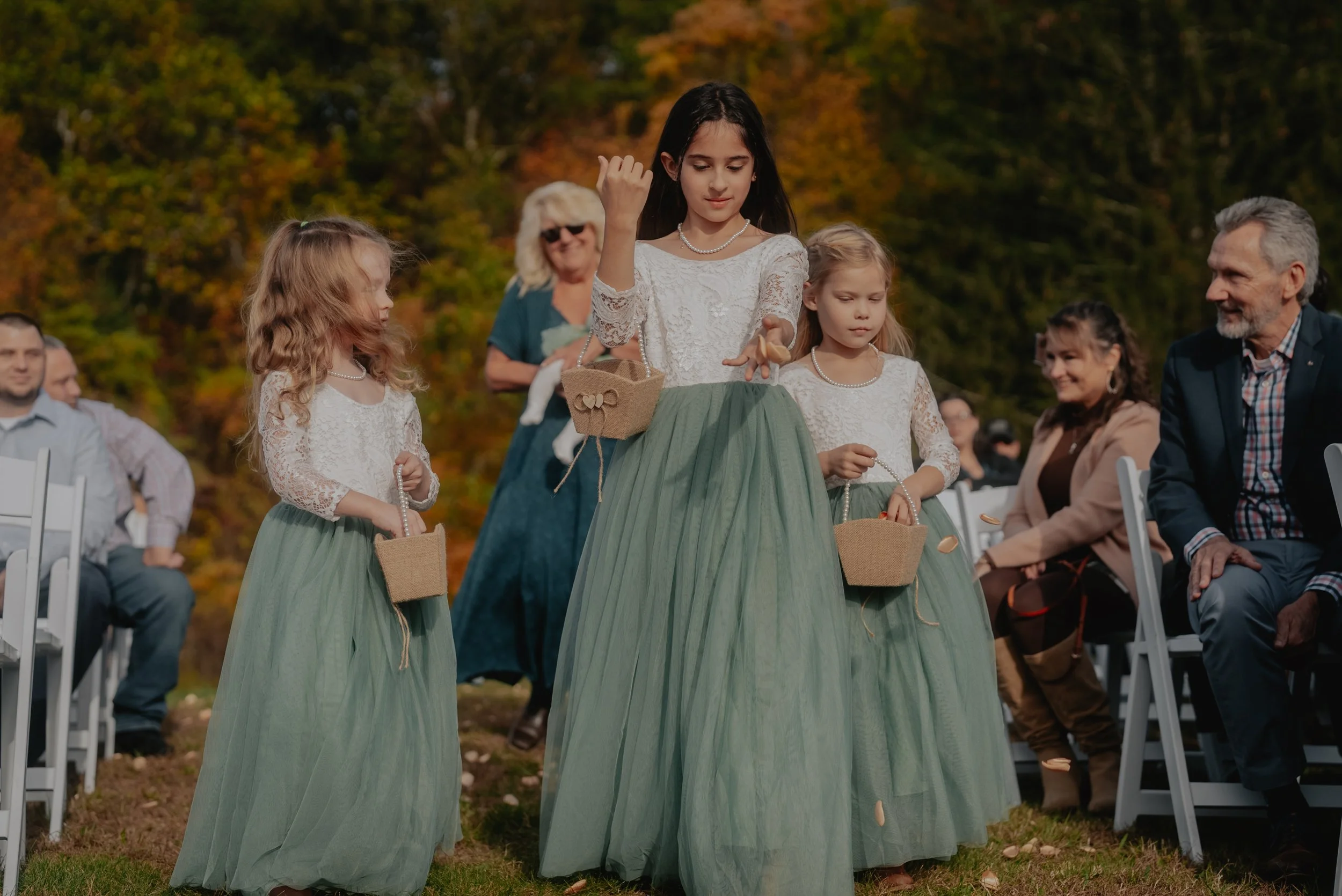 Four young girls dressed in white lace tops and green skirts carry small baskets, participating in an outdoor event amid seated adults.