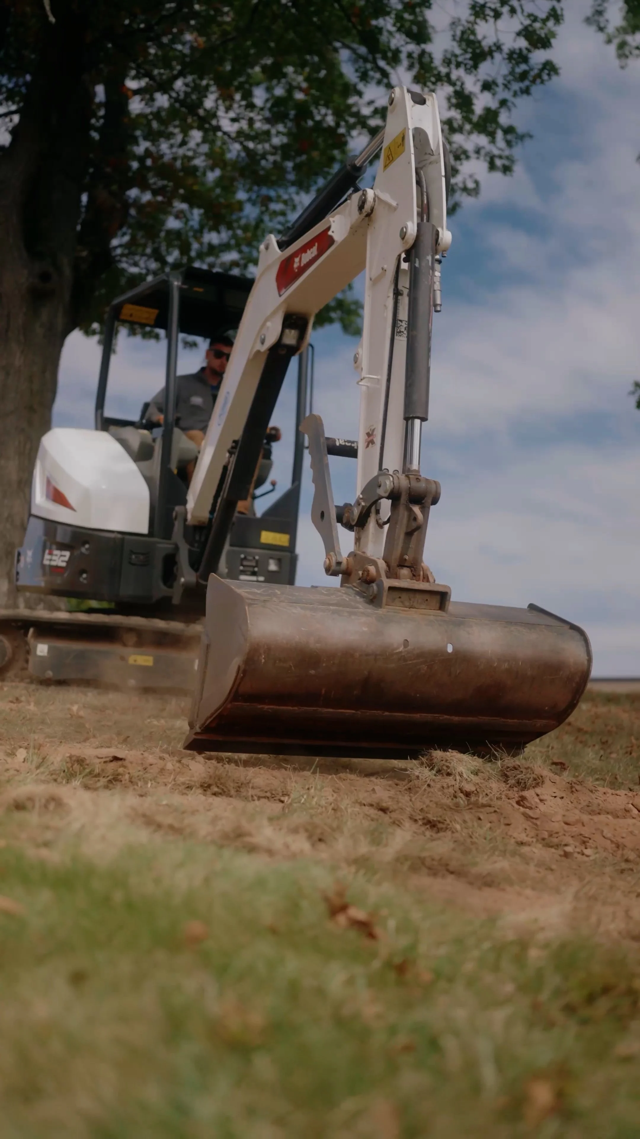Close-up of a small excavator bucket digging into the ground with a person operating it in the background under a tree and blue sky.