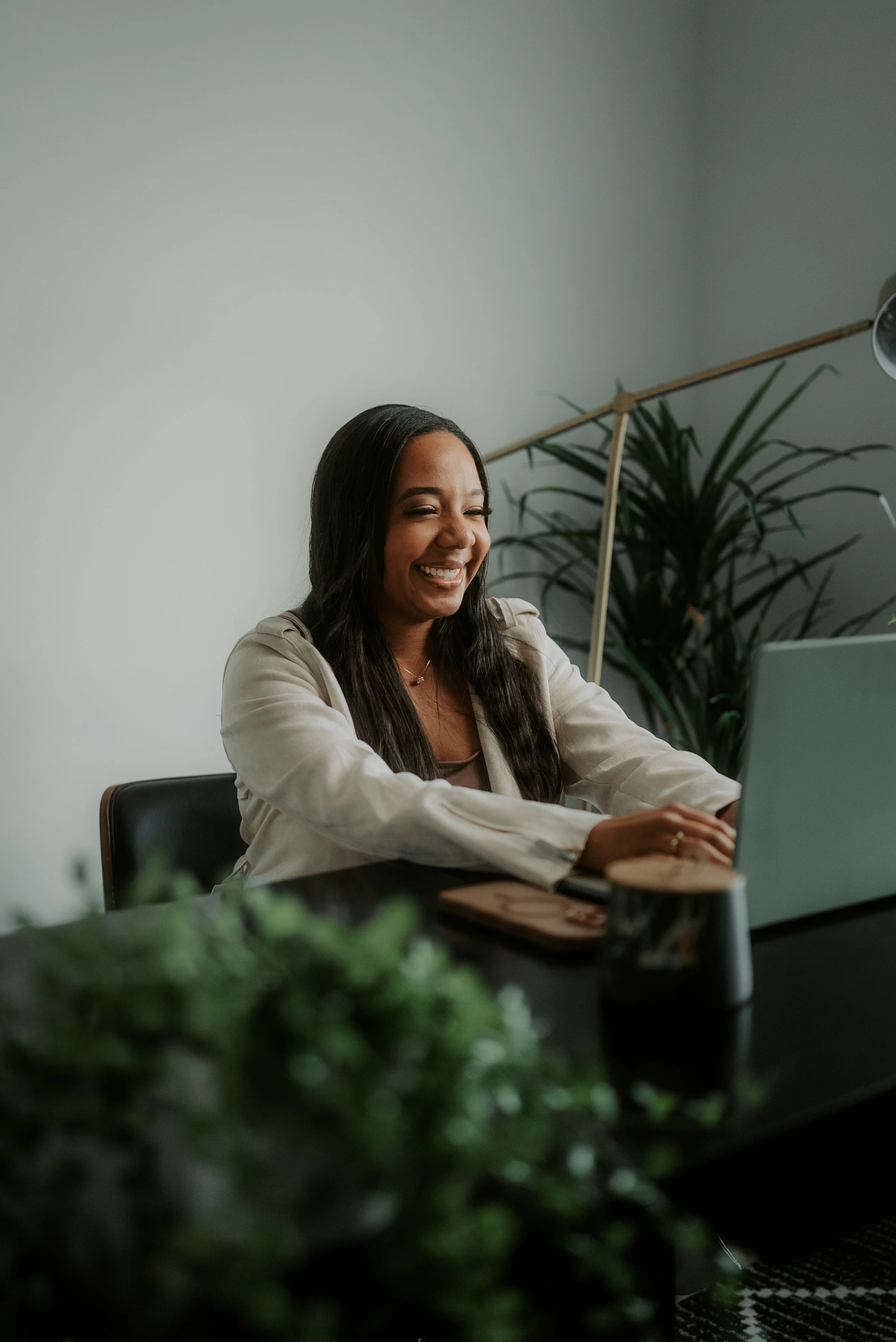 A woman with long dark hair sitting at a desk, smiling while looking at her laptop.