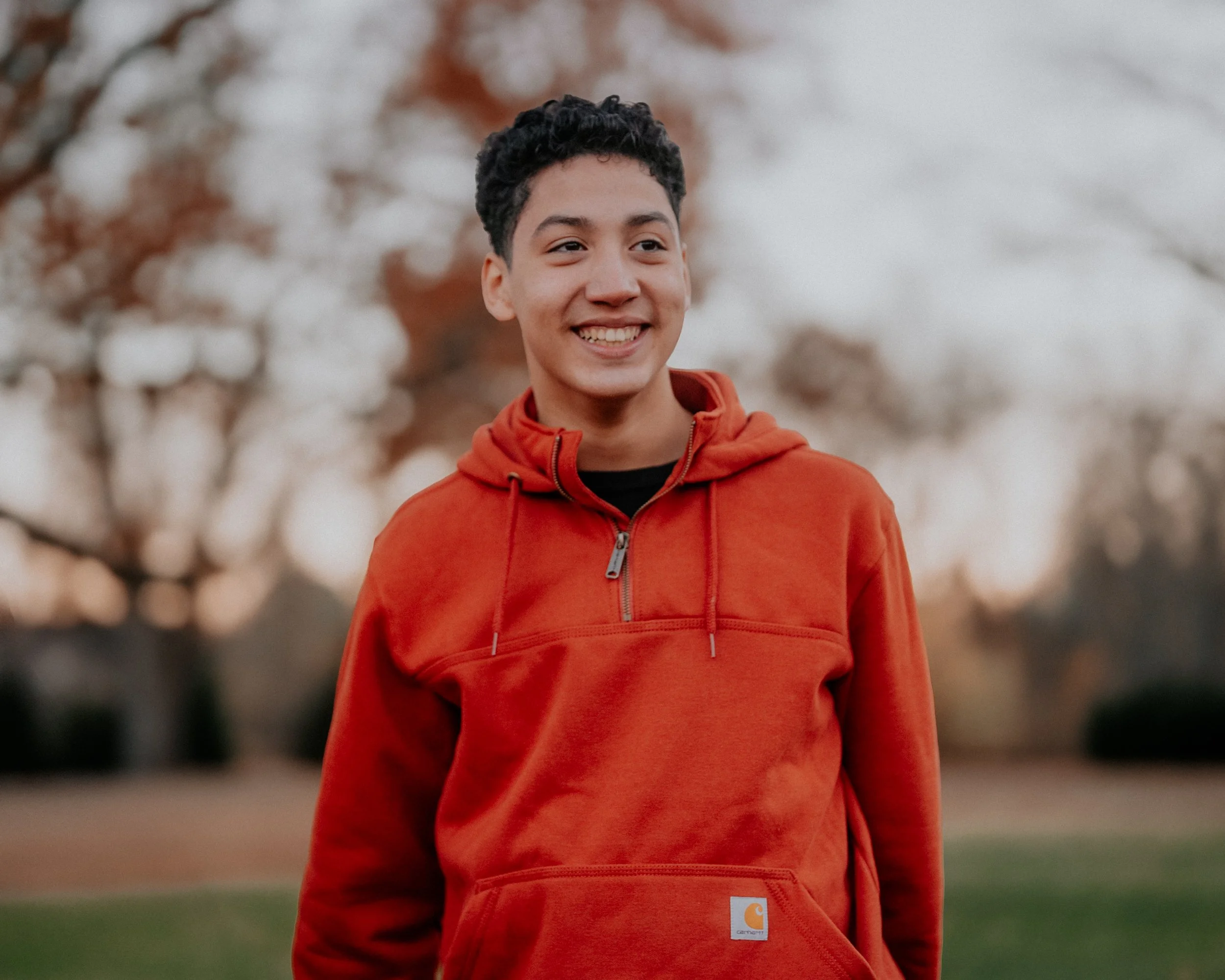A young man smiling outdoors, wearing an orange hoodie, with blurred trees in the background.