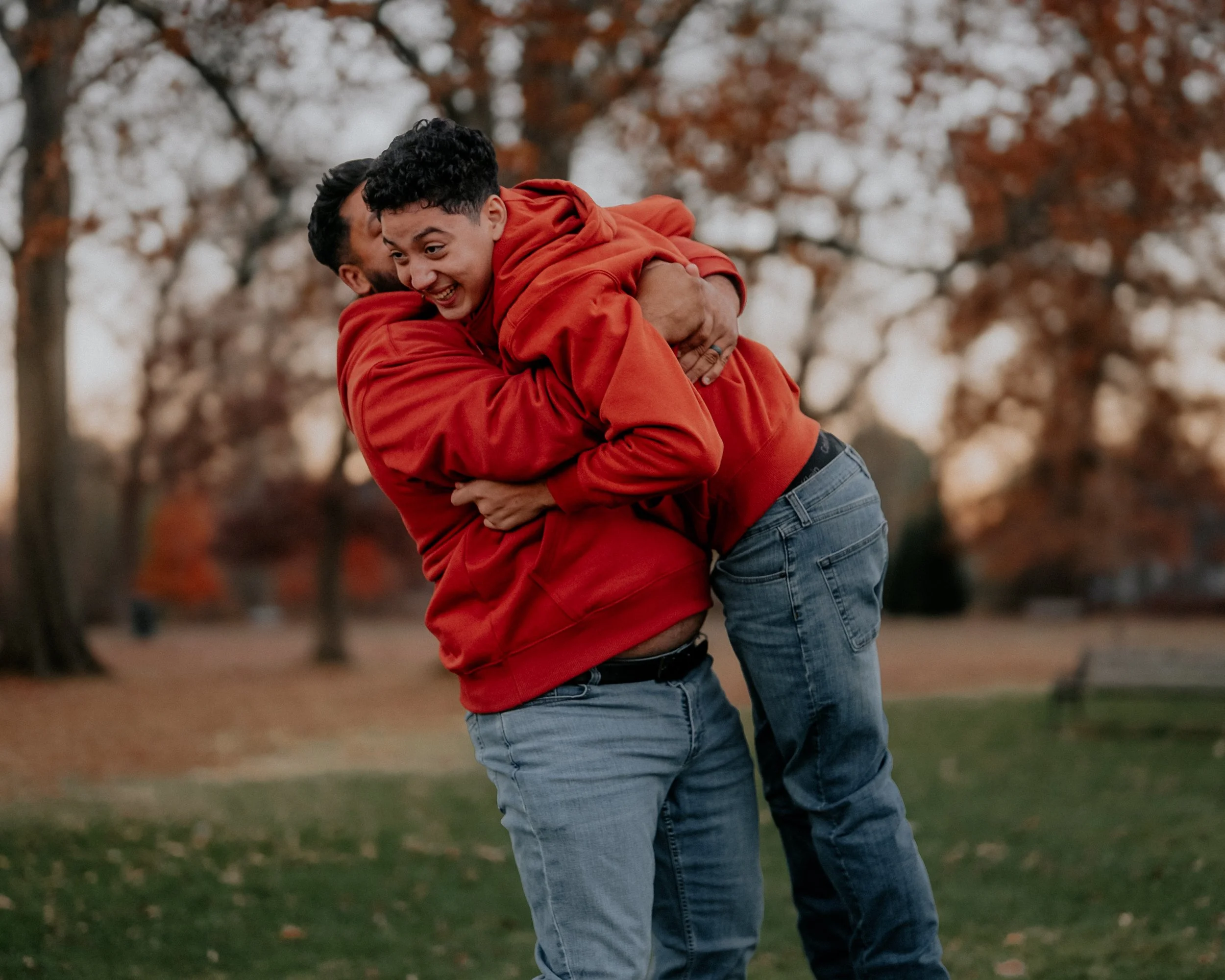 Two young men in red hoodies and jeans are playing together in a park during autumn, with trees and fallen leaves in the background.