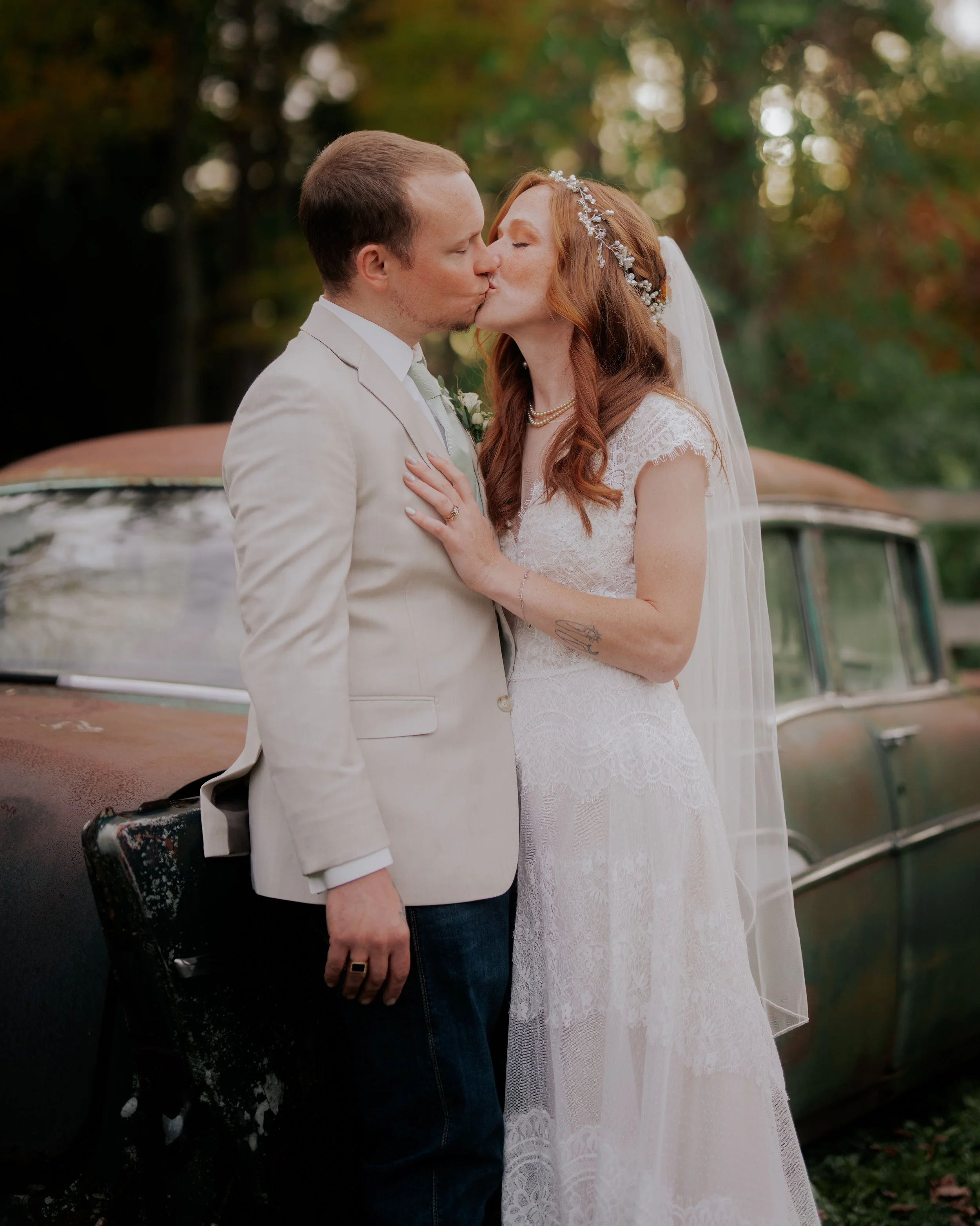 A bride and groom kissing in front of an old rusty car outdoors during sunset.