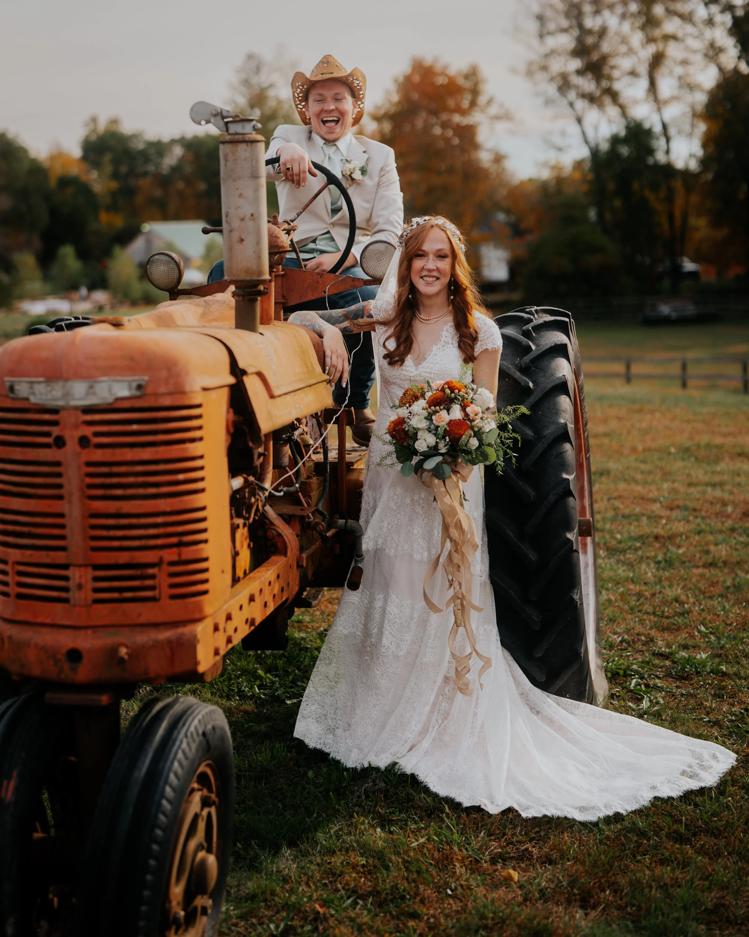 A bride in a white wedding dress holding a bouquet, standing outdoors beside an orange tractor with a man in a cowboy hat sitting on it, during sunset in a rural setting.