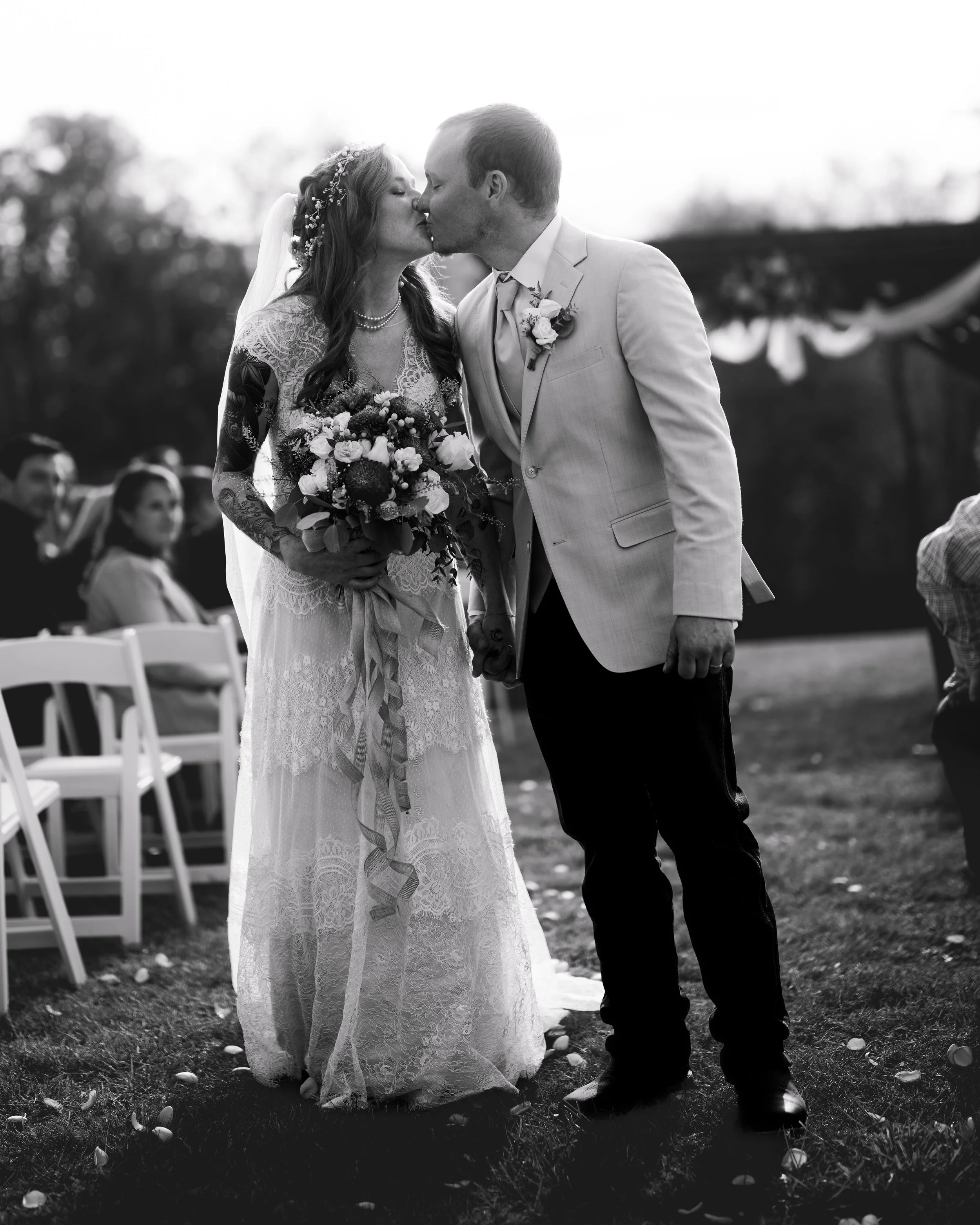 A black-and-white photo of a bride and groom kissing during their wedding ceremony outdoors, with wedding guests seated in the background.