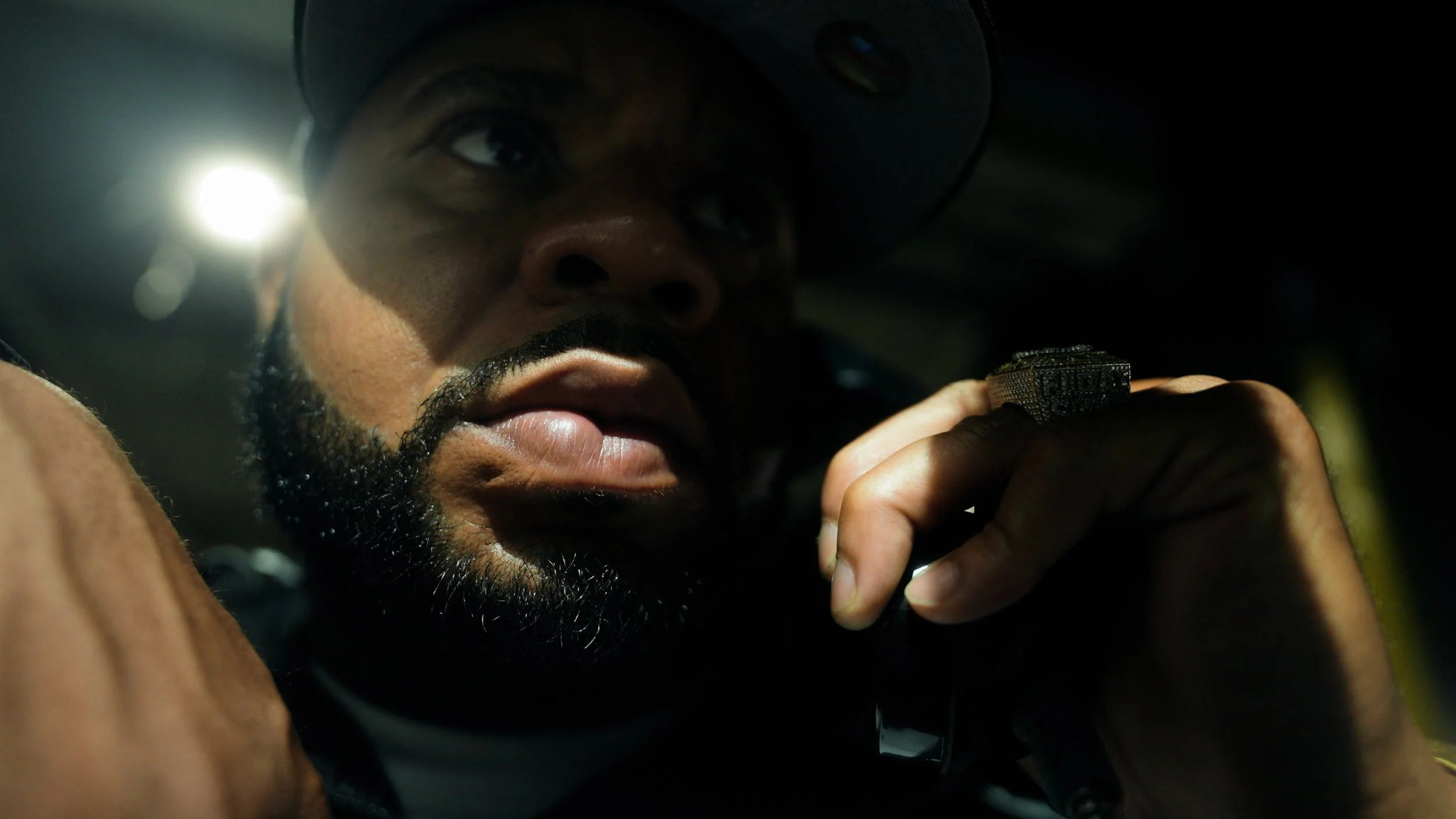 Close-up of a man with a beard wearing a hat, holding a phone to his ear, with a dark background and a bright light in the distance.
