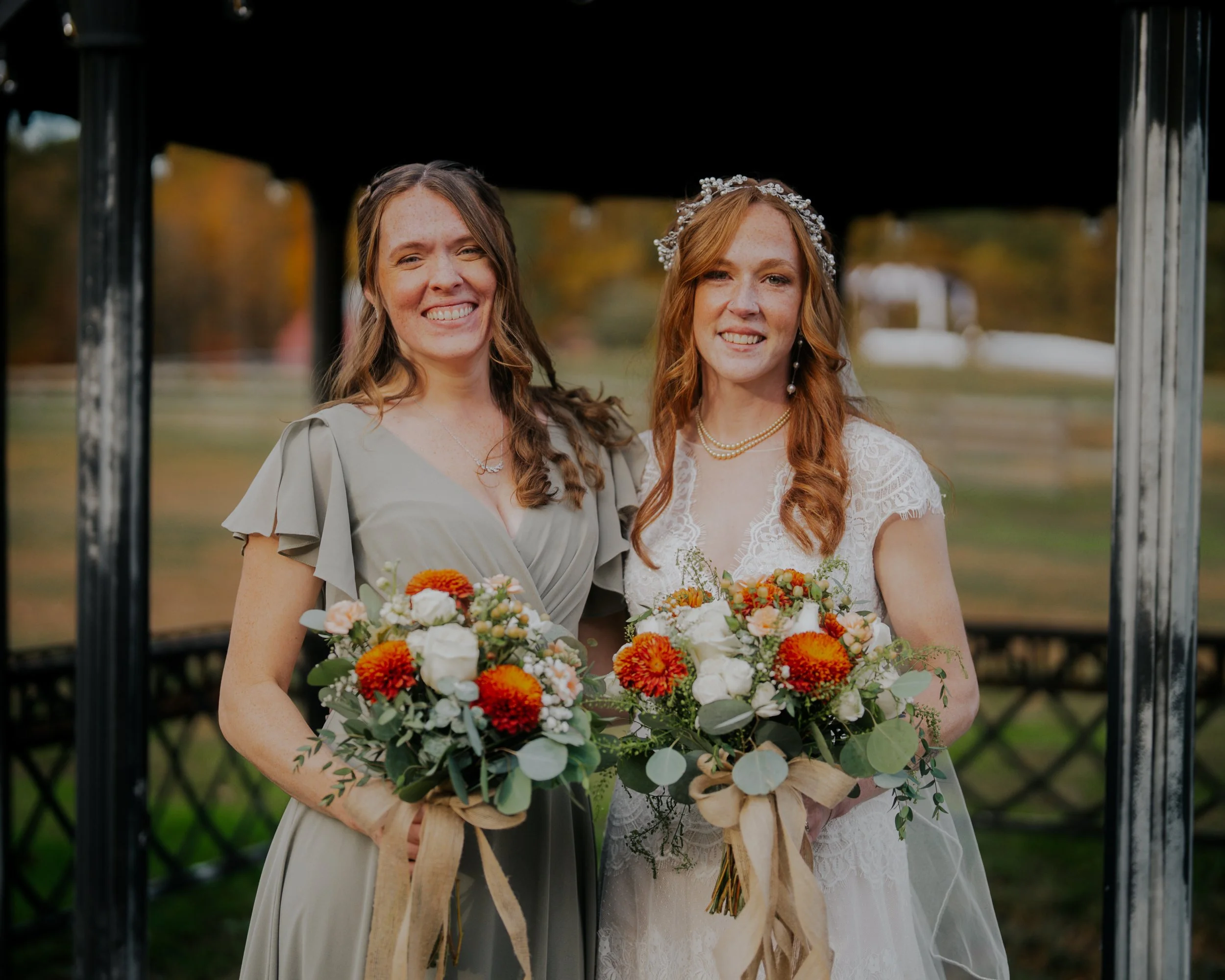 Two women standing together outdoors during daytime, holding bouquets of flowers, smiling, one wearing a white wedding dress with lace, the other in a light gray dress with ruffled sleeves.