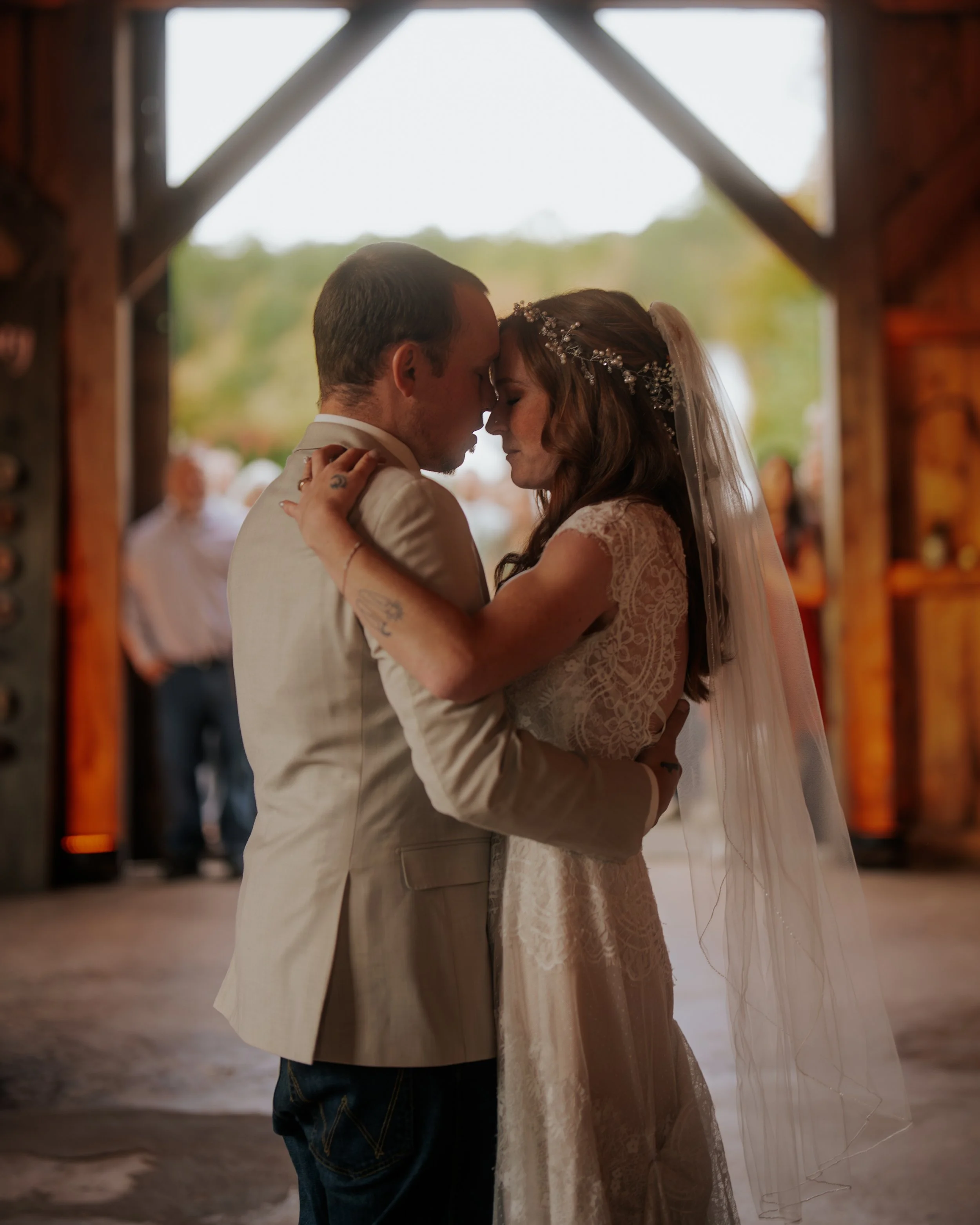 A bride and groom embrace during their wedding inside a rustic barn, with an outdoor scene visible through the open barn doors in the background.