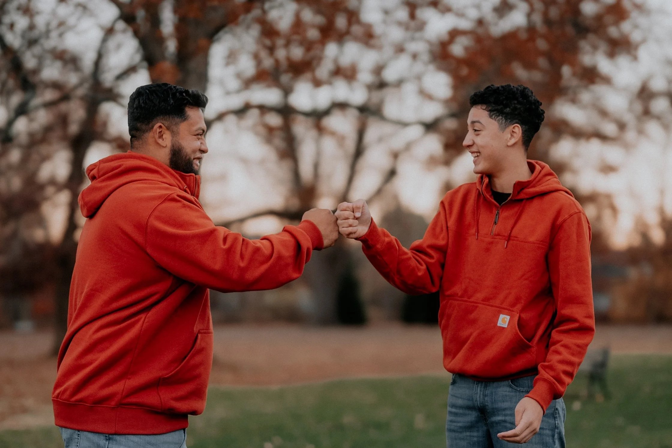 Two young men wearing orange hoodies are greeting each other with a fist bump outdoors, with trees in the background.