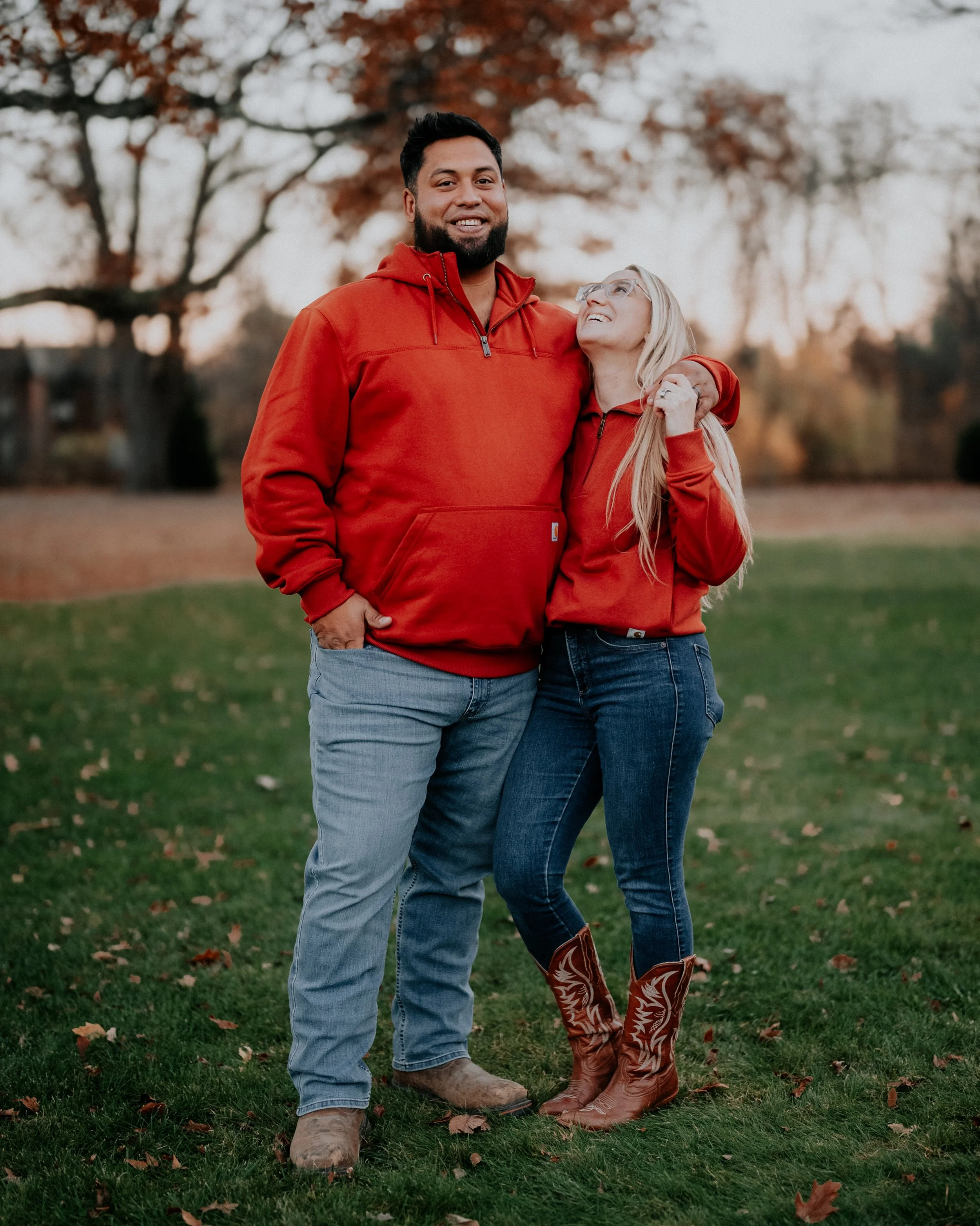 A man and a woman wearing red hoodies standing outdoors on grass with trees in the background during fall; the woman is looking up at the man with a smile.