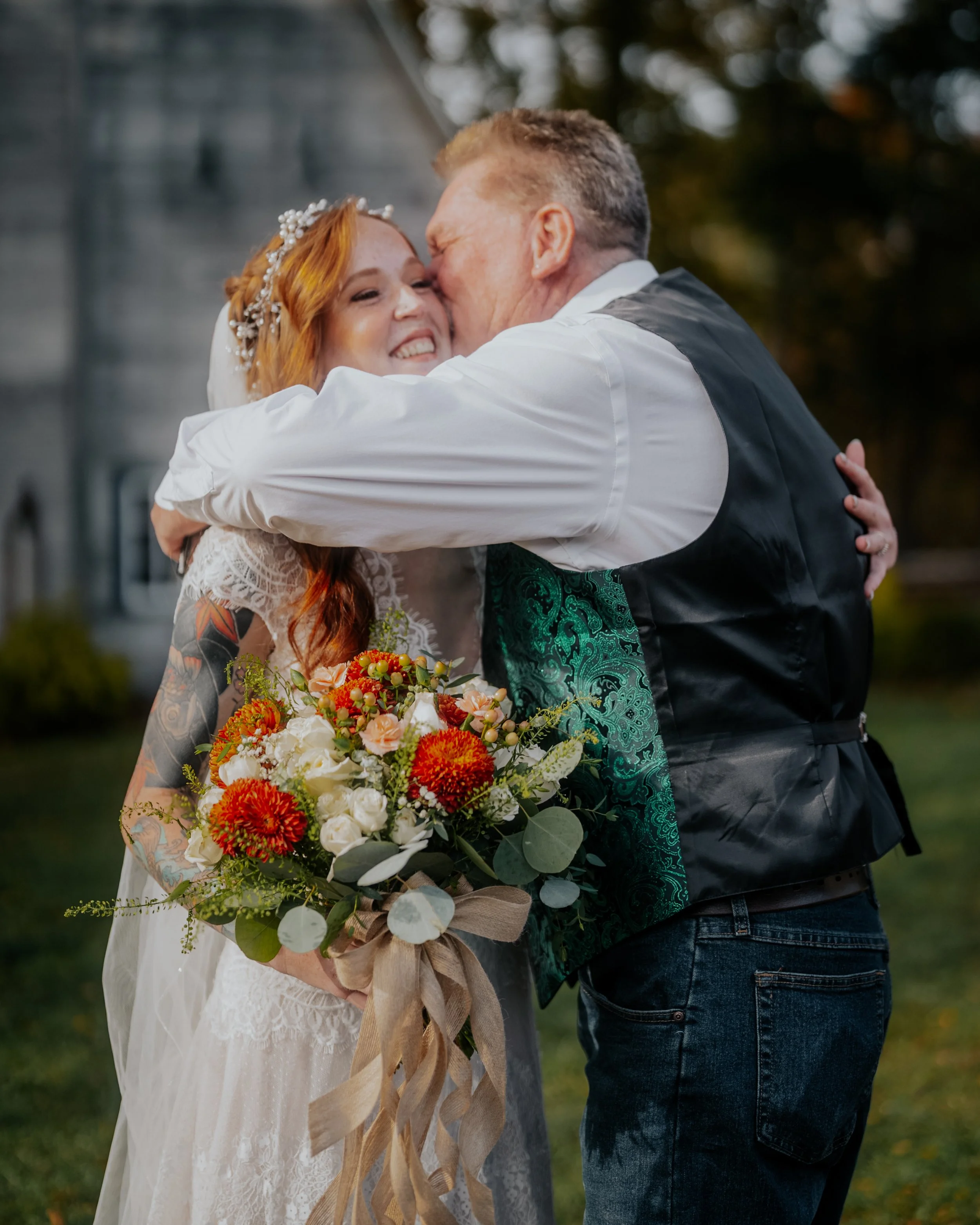A bride with long red hair and tattoos on her arm, holding a bouquet of flowers, hugging and kissing a man in a black vest, outside in a garden area.