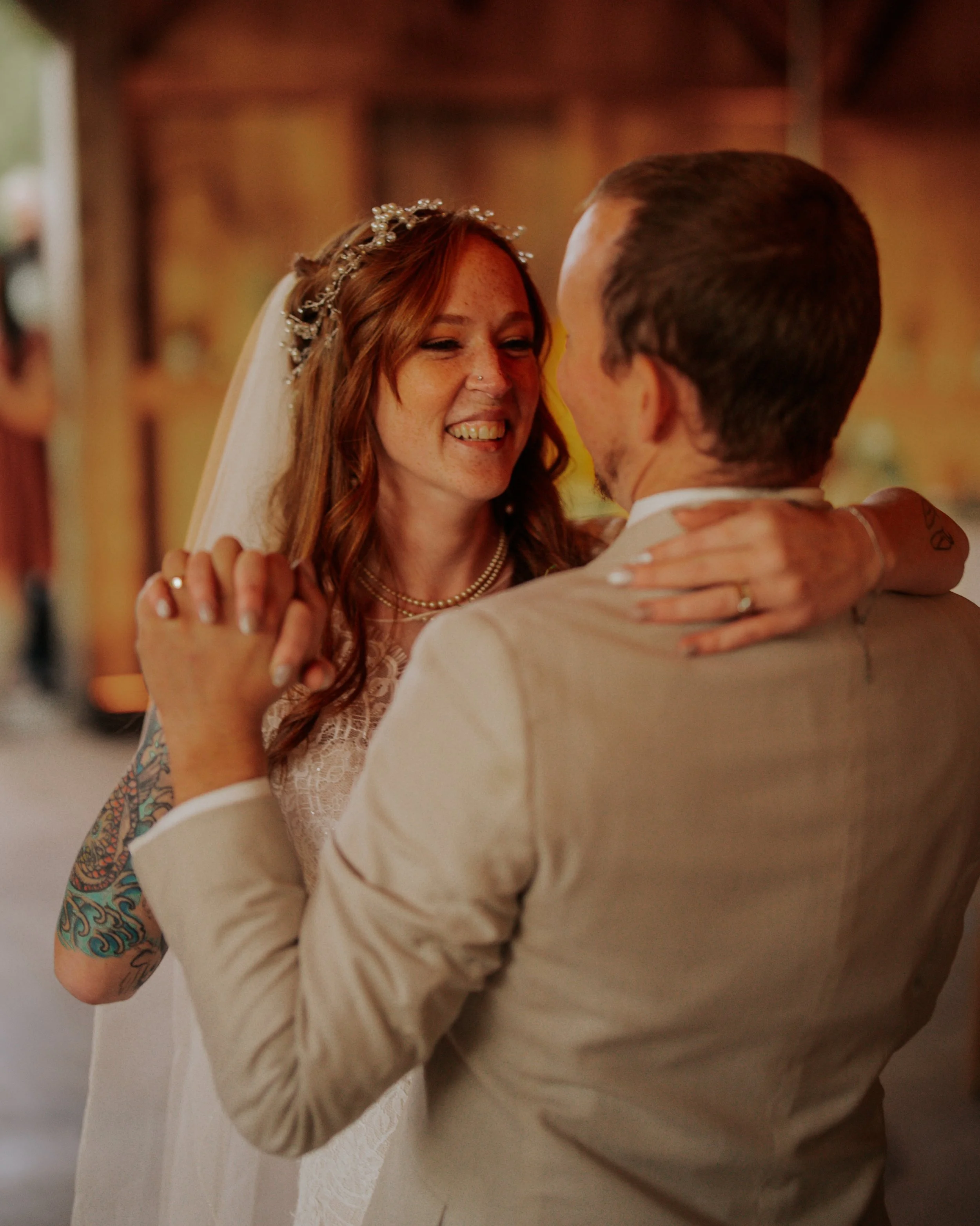 A bride and groom dance closely during their wedding reception, smiling at each other with warm lighting and a rustic background.