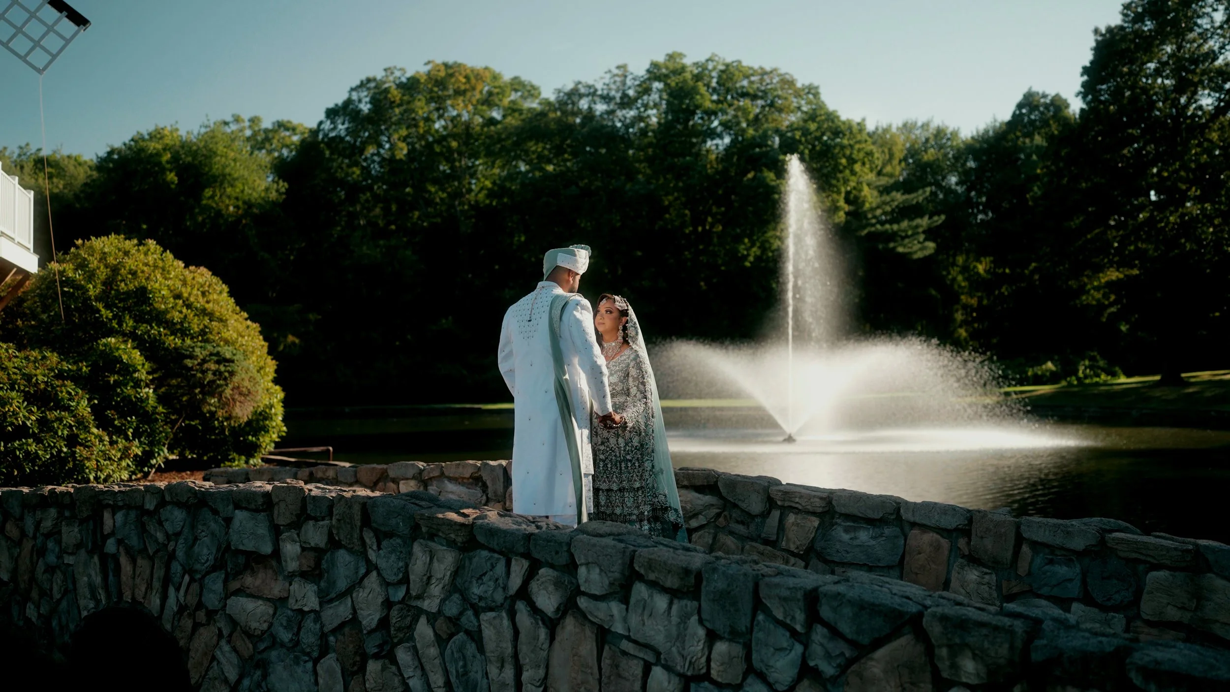 A bride and groom holding hands and looking into each other's eyes near a pond with a water fountain, surrounded by greenery and a stone wall.