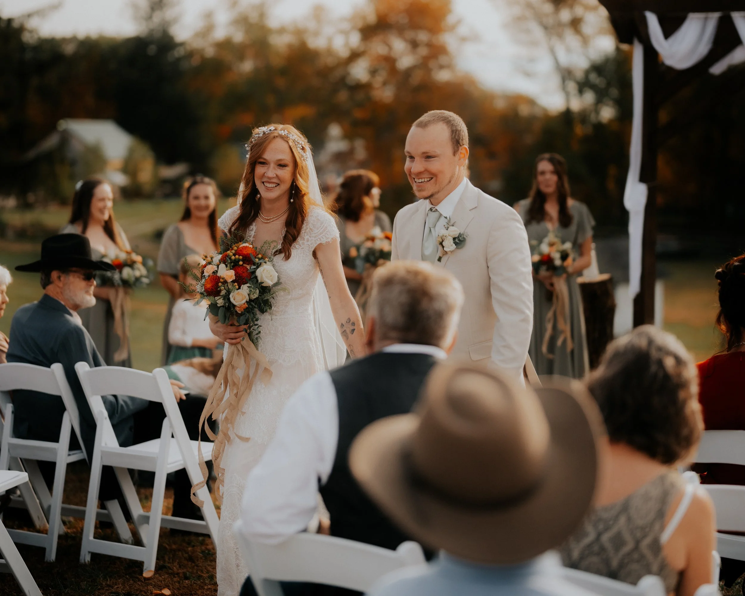 A bride and groom smiling at their wedding ceremony outdoors during sunset, with bridesmaids and guests seated nearby.