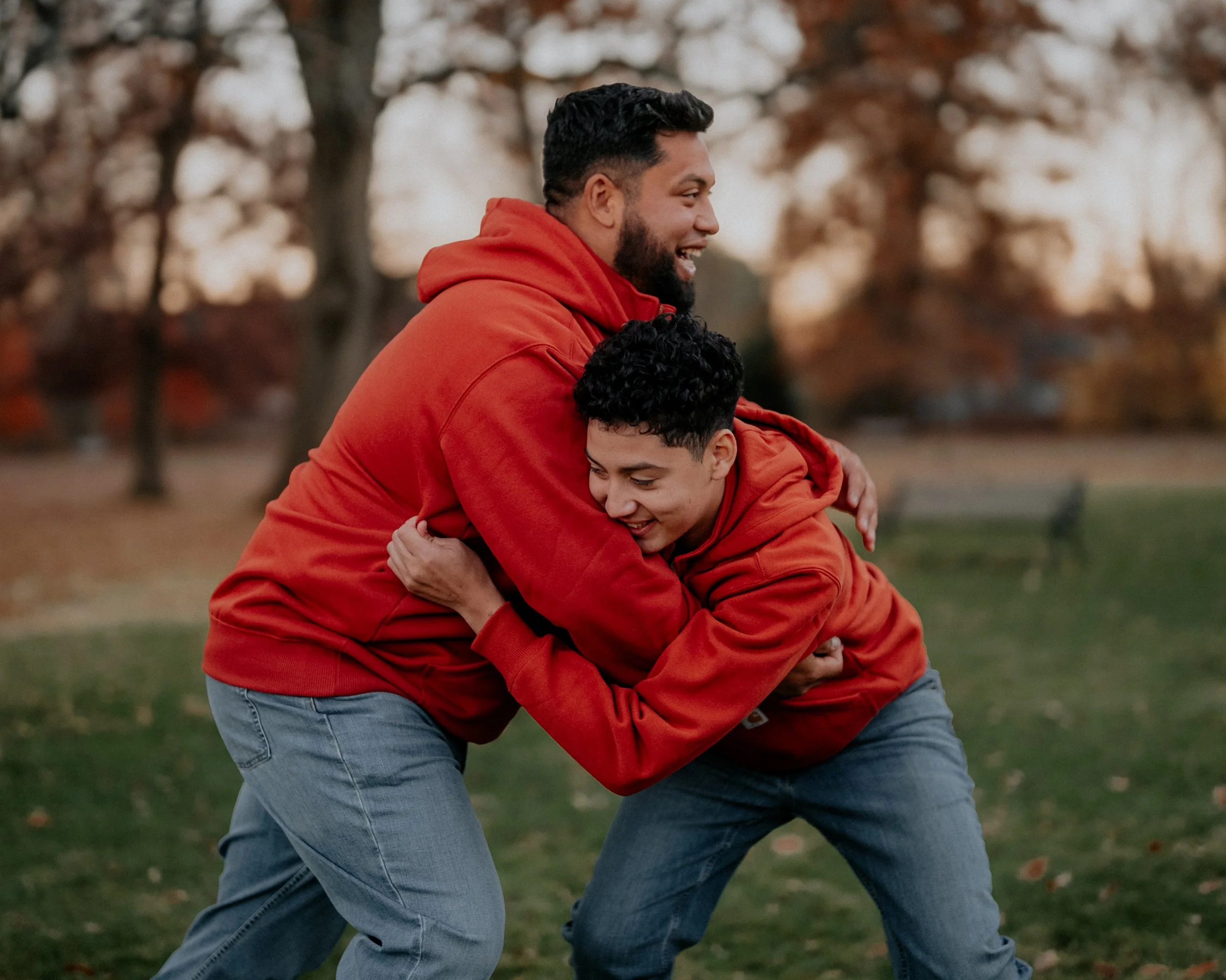 Two males wearing red hoodies are playfully wrestling outdoors in a park during autumn, with trees and fallen leaves in the background.
