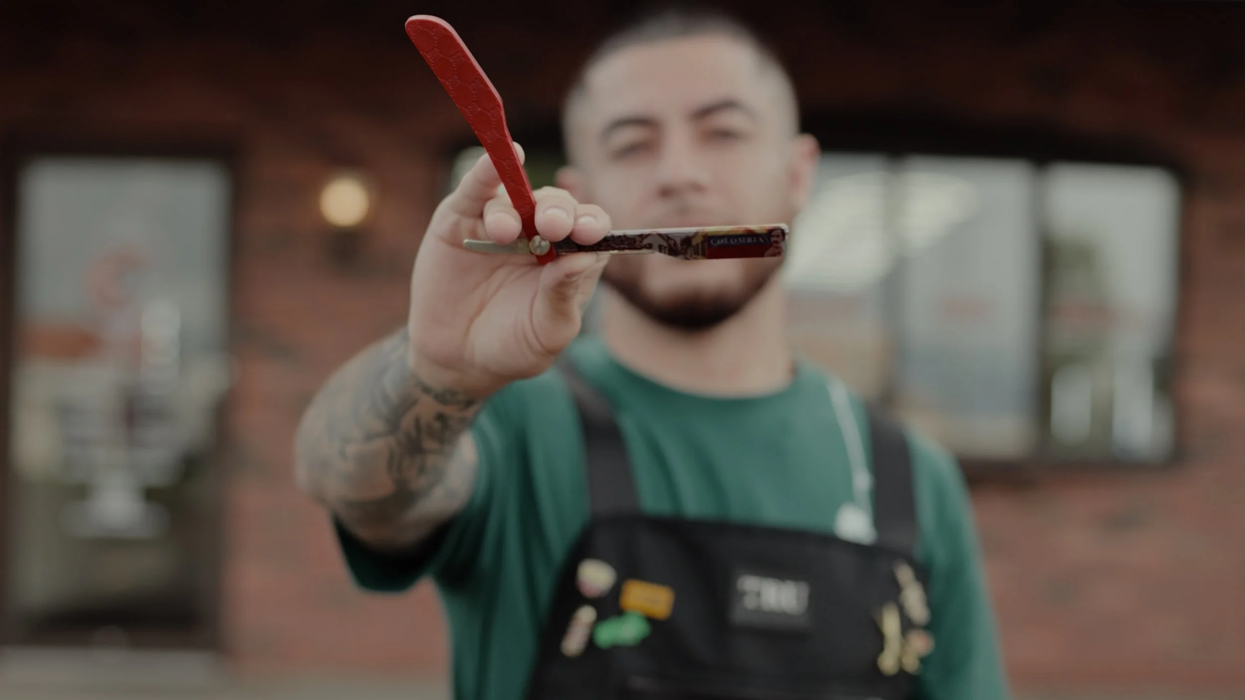A man with tattoos on his arm, wearing a green shirt and black apron, holds sunglasses towards the camera with the background of a brick building.