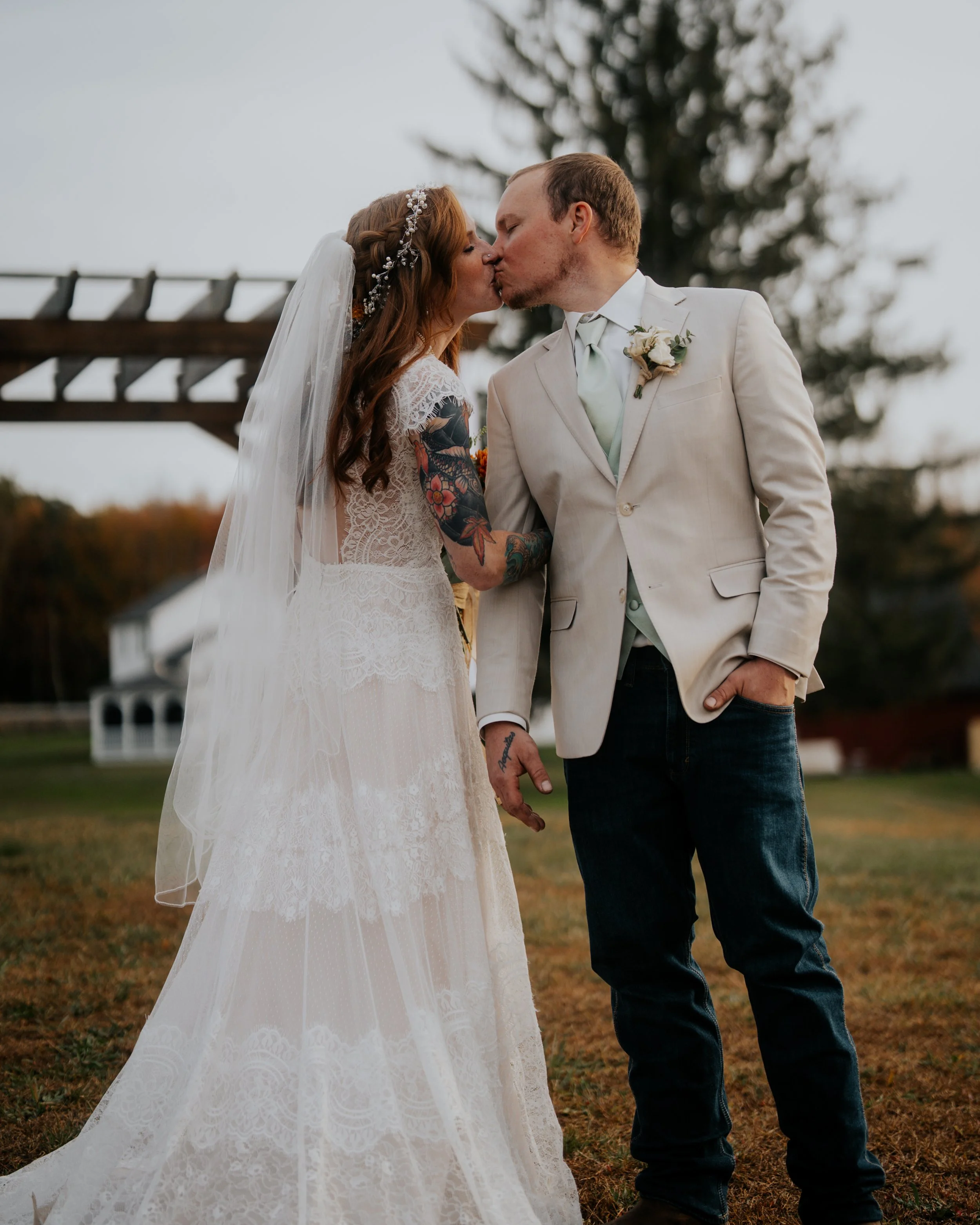 A bride and groom kiss outdoors during their wedding ceremony. The bride has long red hair, tattoos on her arm, and wears a lace wedding dress with a veil. The groom has short hair, wears a light-colored suit jacket, tie, and dark jeans, with a bouto