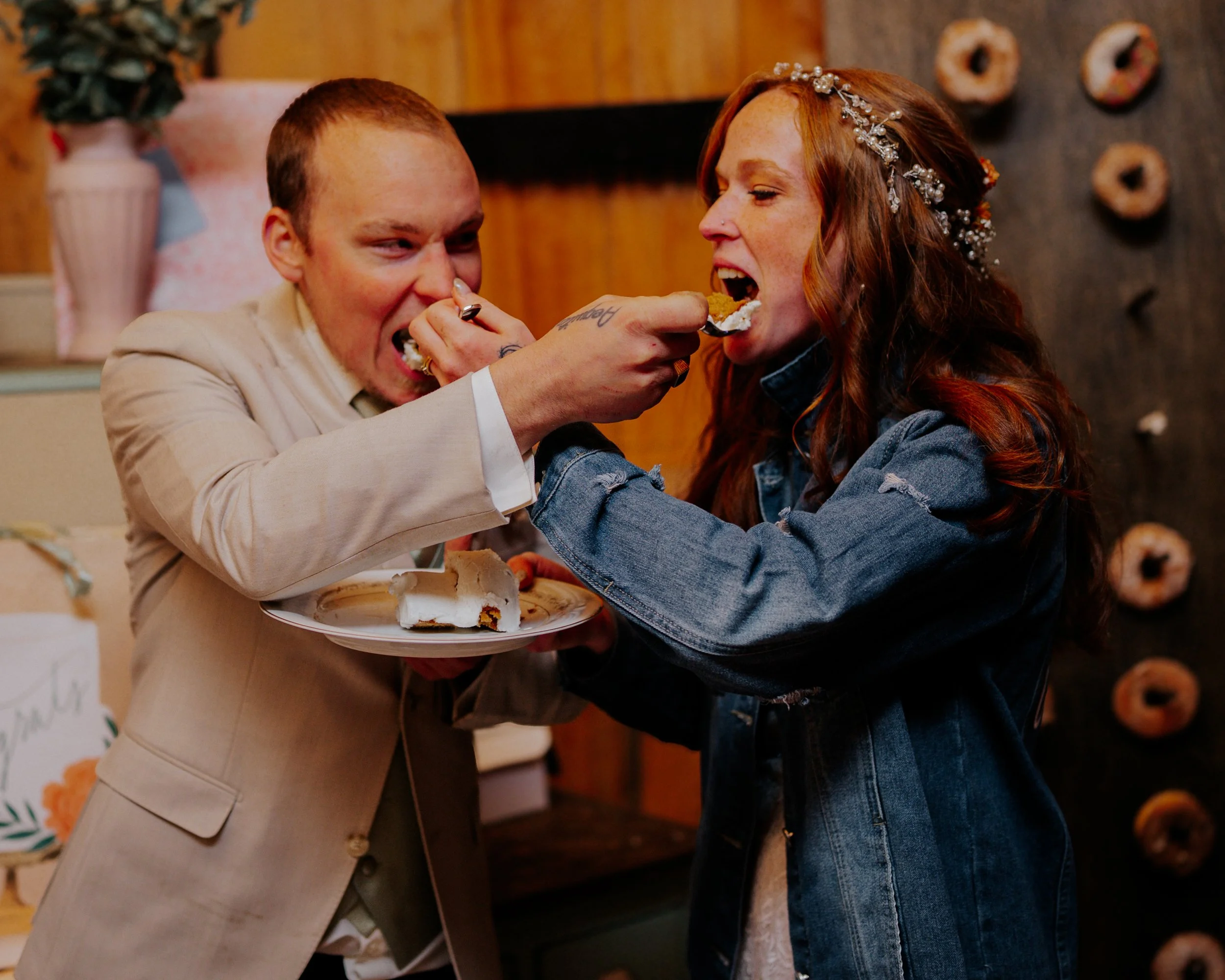 A man in a beige suit feeding a slice of cake to a woman with red hair and a headband, inside a cozy wooden room decorated with donuts on the wall.