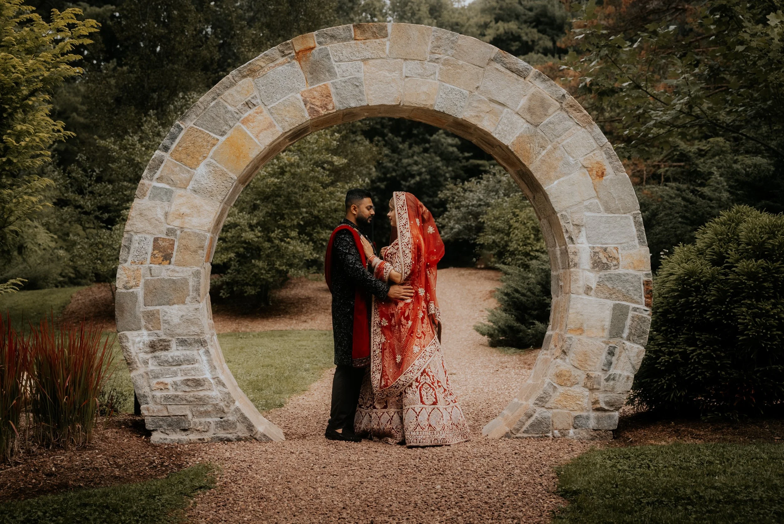 A couple in traditional South Asian wedding attire standing closely under a stone archway in a garden setting, with green bushes and trees in the background.