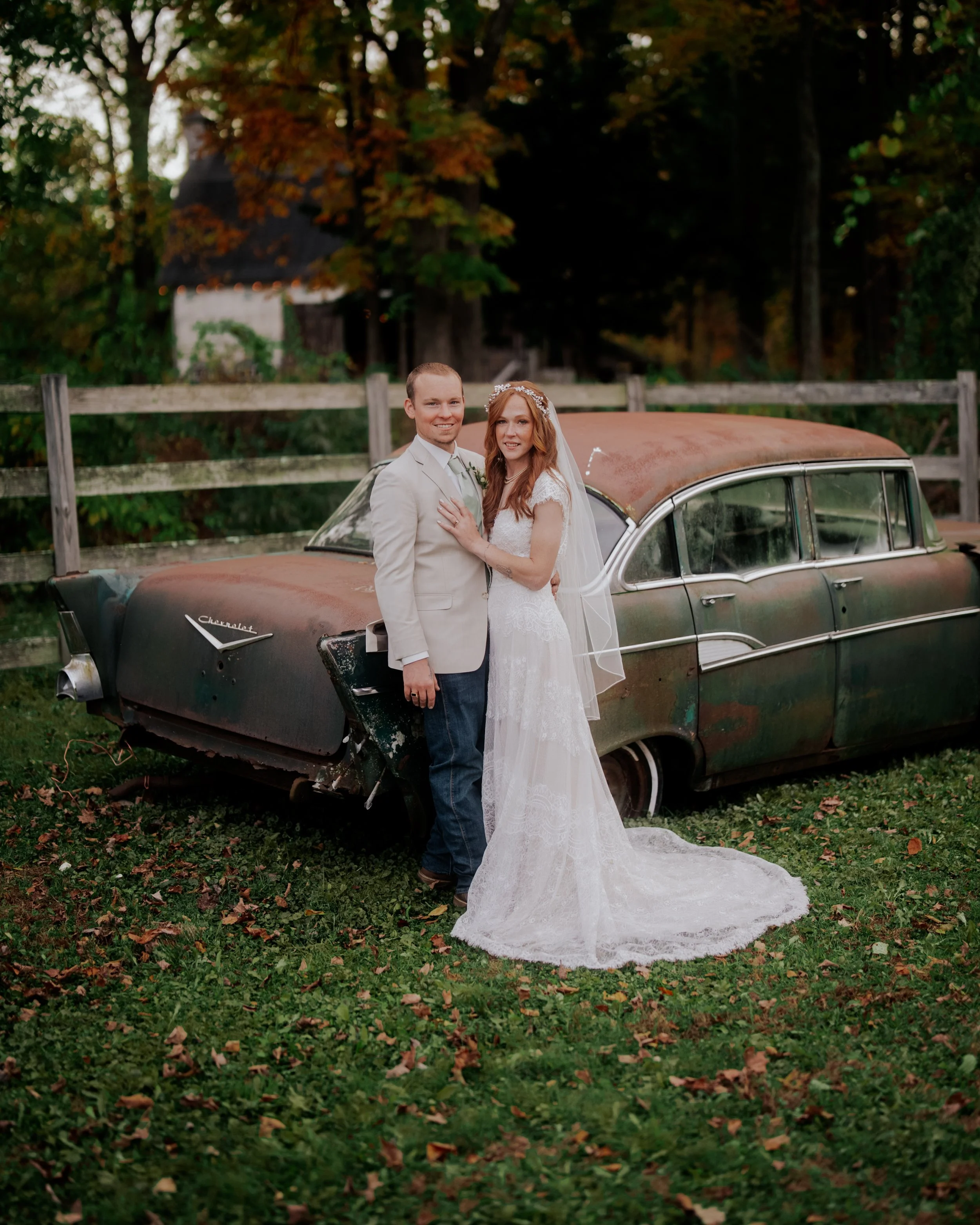 A newlywed couple stands in front of a rusty old car outdoors with trees and a wooden fence in the background. The groom wears a light-colored suit and the bride wears a white lace wedding dress with a veil.