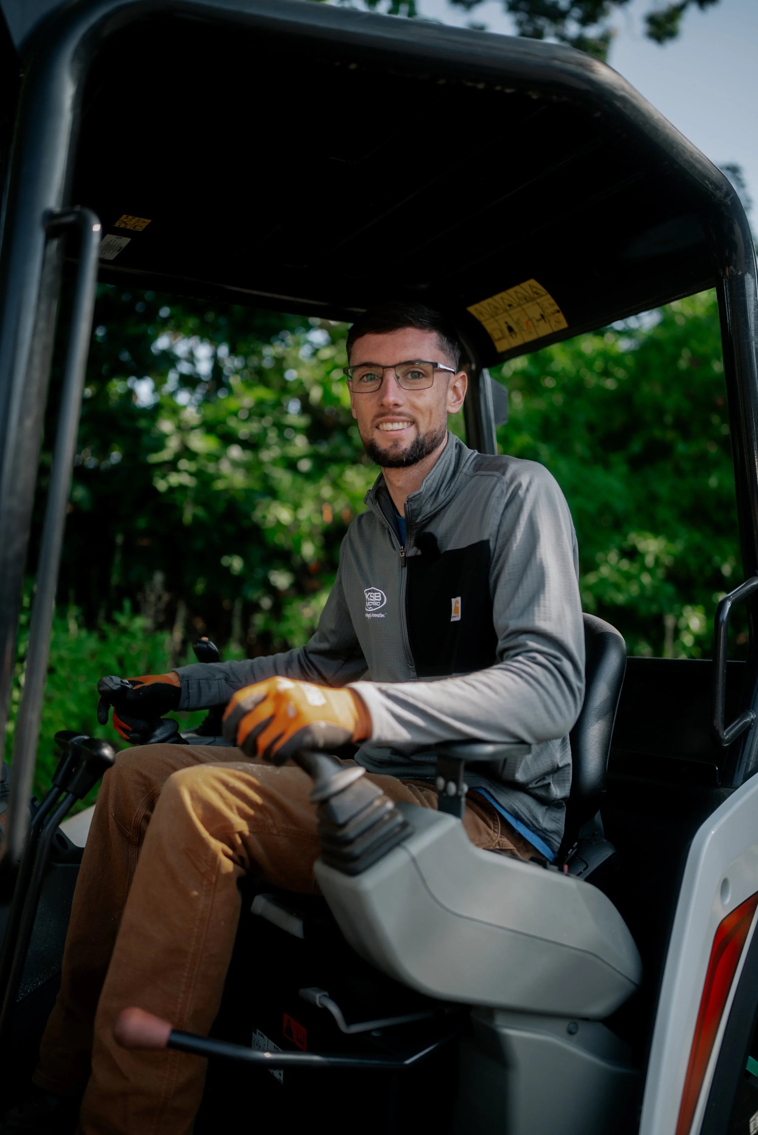A man wearing glasses, gloves, and a gray jacket sitting inside a tractor, smiling at the camera, with green trees in the background.