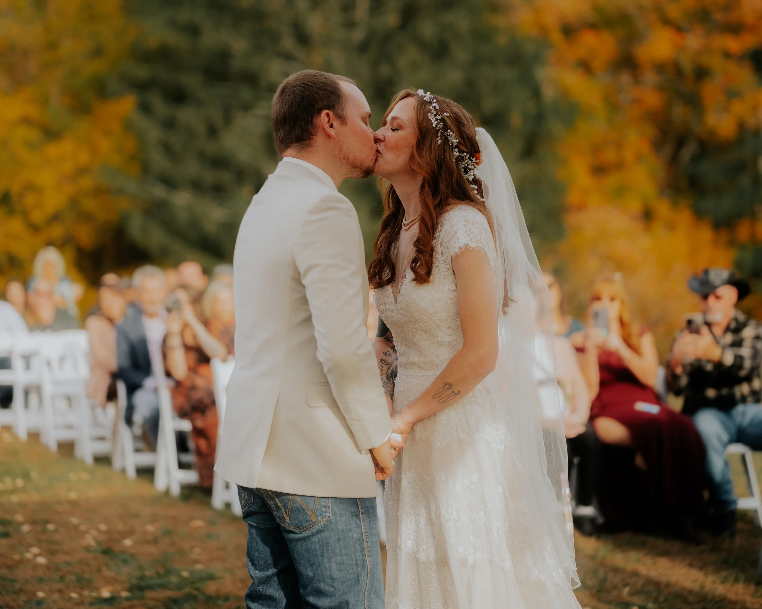 A bride and groom kiss during an outdoor wedding ceremony in autumn, with seated guests in the background.